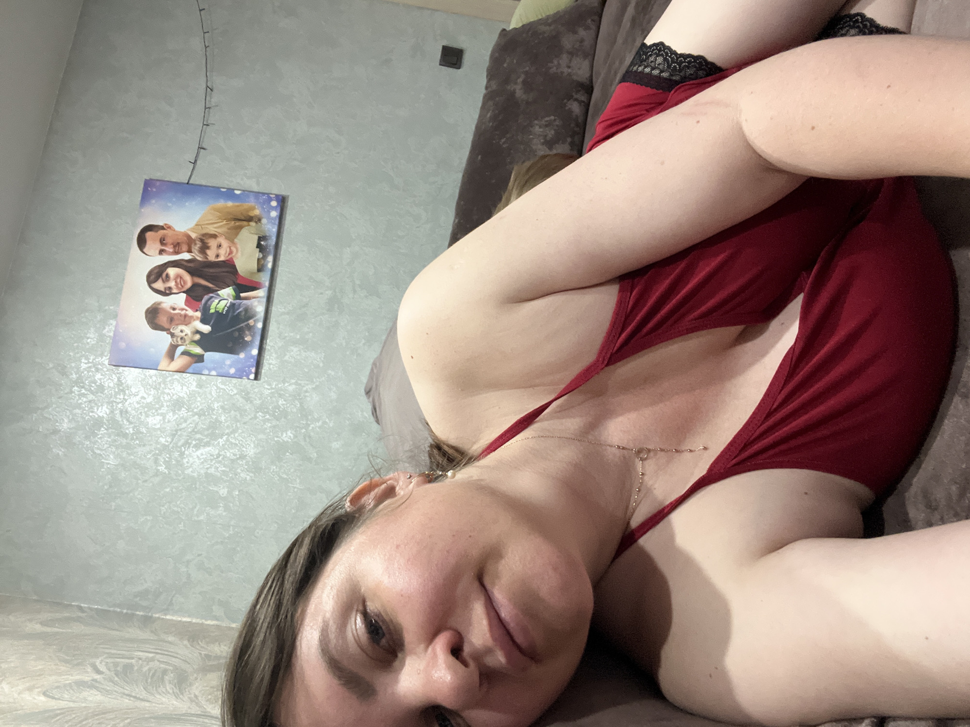 A woman with medium-length brown hair, wearing a red top with black lace trim, is lying on a couch. She is looking towards the camera. On the wall behind her, a framed picture depicts a family of four. The environment appears to be an indoor setting, possibly a living room, with a textured light-colored wall and a dark-colored couch. The scene does not provide enough visual information to determine the specific location within the city of Noviy, Russia.