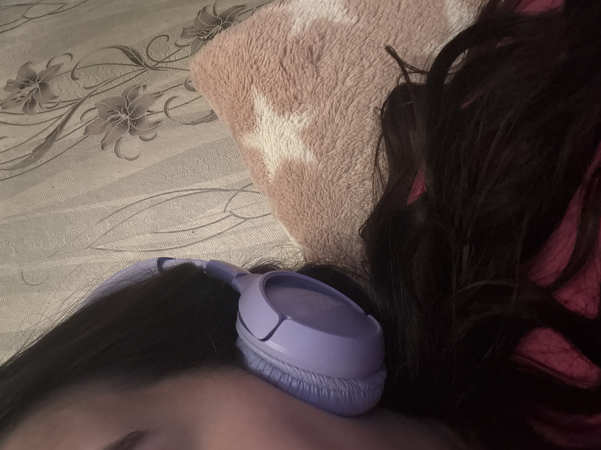 A close-up, eye-level shot shows a person with dark hair and a lavender-colored pair of headphones resting on their head. A pink, fuzzy pillow is partially visible behind the person's head. The background is a patterned surface that appears to be a bed sheet or blanket. No information within the image indicates location in Dobrovnitsa, Bulgaria.