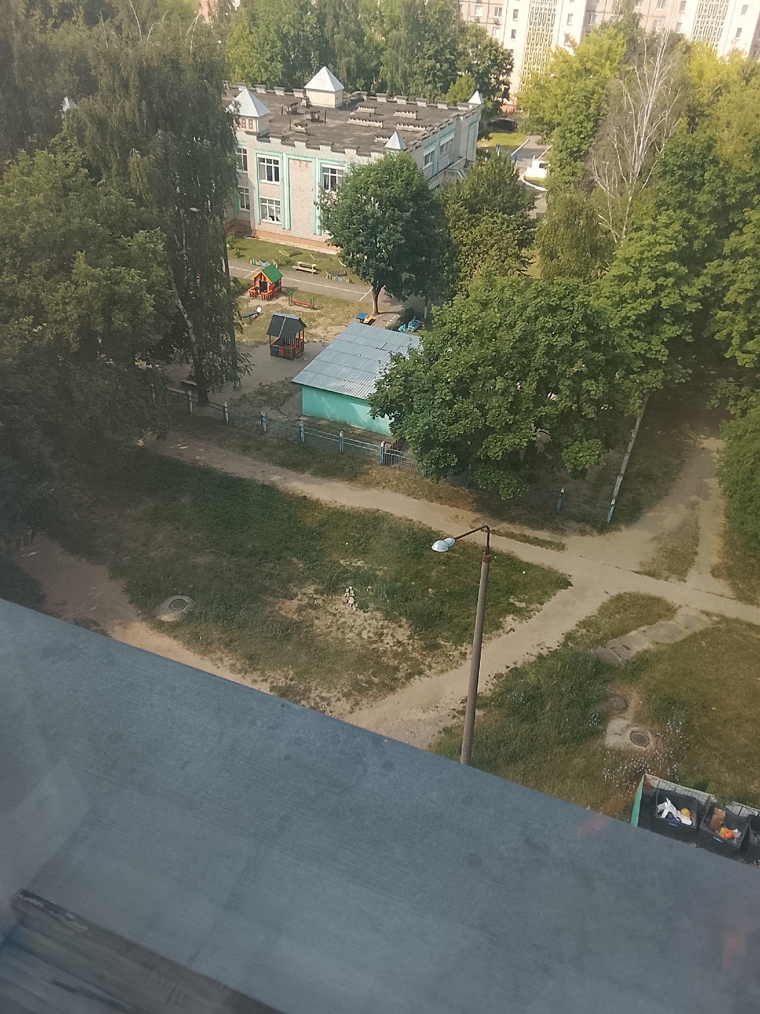 The image is an aerial view of a playground area next to a building, likely a kindergarten or school, in the city of Zhlobin, Belarus. The building has a light-colored brick facade and a grey roof with decorative turrets. Adjacent to the building is a playground with a small, colorful playhouse and other playground equipment. A fence separates the playground from a dirt path and grassy areas. A street lamp with a single light is visible in the lower right quadrant of the image. In the far background, several tall apartment buildings can be seen amidst lush green trees. A row of garbage bins is also present near the bottom right corner of the image, some of which contain visible trash. The overall scene depicts a typical urban environment in Belarus, with a focus on a recreational space for children.