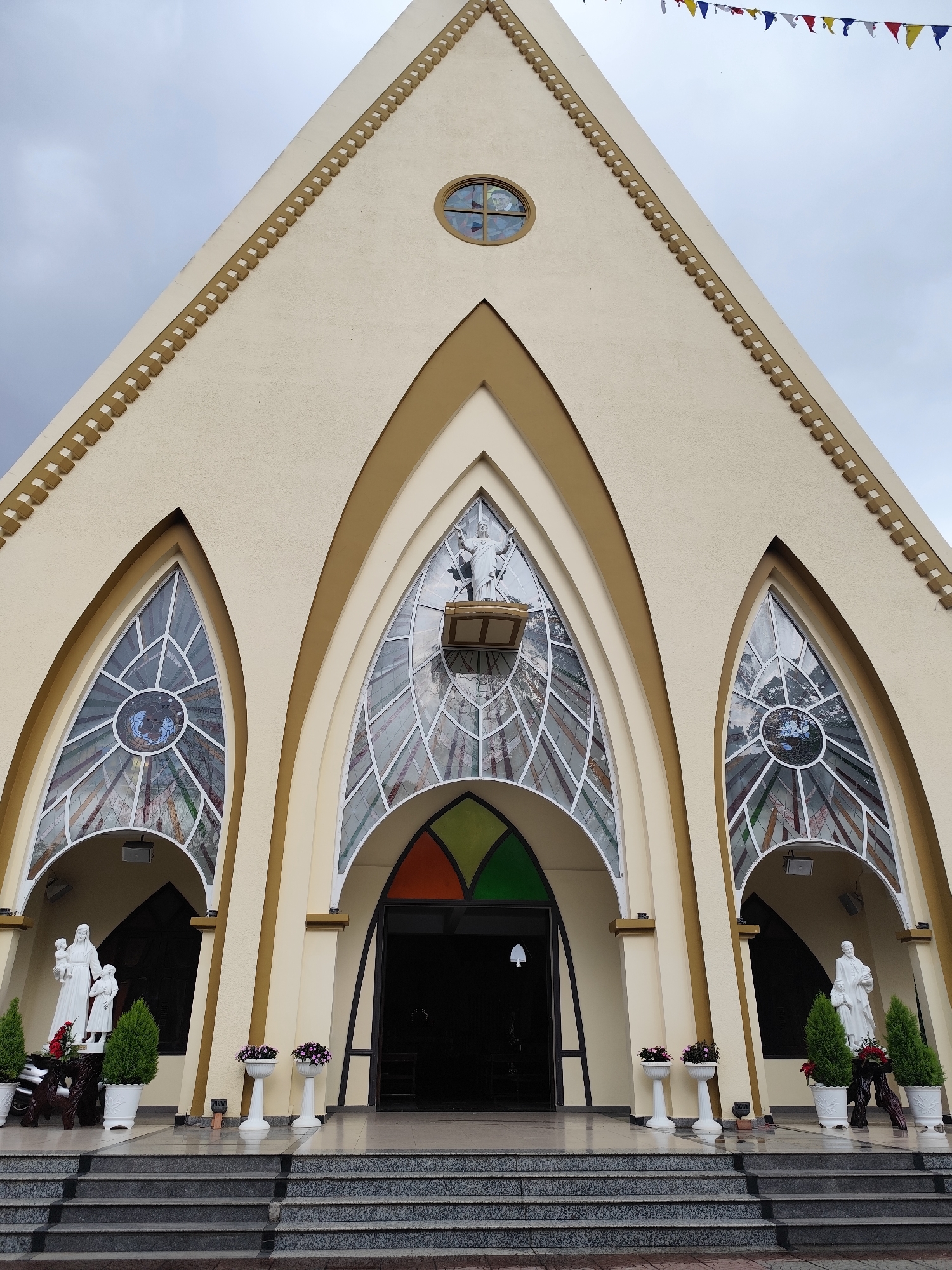 The image depicts the facade of the Da Lat Cathedral, located in Da Lat, Vietnam. The building features an A-frame design with three arched entryways. The central arch is larger and houses the main doors. Stained glass windows are visible within the arches. Statues flank the side entryways. The building is painted in neutral tones with darker trim. Steps lead up to the entrance. Decorative flower pots are placed along the steps and in front of the building. A string of colorful flags hangs above the building. The sky is overcast.