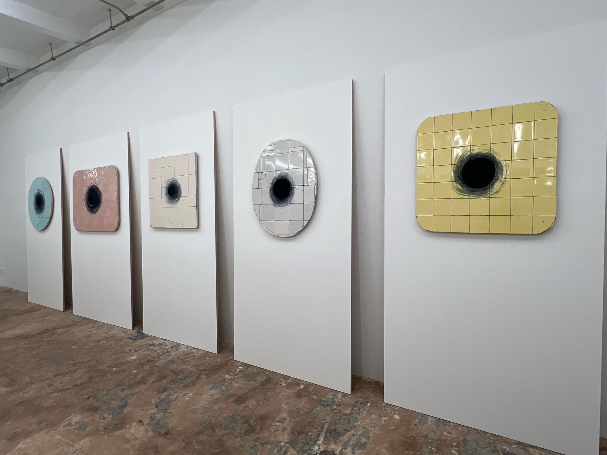 A row of five art pieces is displayed on white panels against a white wall in what appears to be an art gallery. The floor is a rough, concrete-like material with areas of discoloration. The art pieces are mounted on individual white panels, each featuring a central, dark circular element. From left to right, the first piece is circular and light blue. The second piece is a rounded square shape in a muted pink hue. The third piece is a square with a light cream-colored tiled surface. The fourth piece is a circular element with a tiled surface, predominantly gray and white. The fifth and final piece is a rounded square shape covered in yellow tiles. All five pieces have a dark, almost black, void-like circle in their center, with some exhibiting brushstroke-like marks radiating outwards. Above the art, conduit piping runs along the ceiling, suggesting an industrial or functional aspect to the gallery space. The overall impression is one of minimalist contemporary art. There are no people visible in the image. The scene is static and silent.