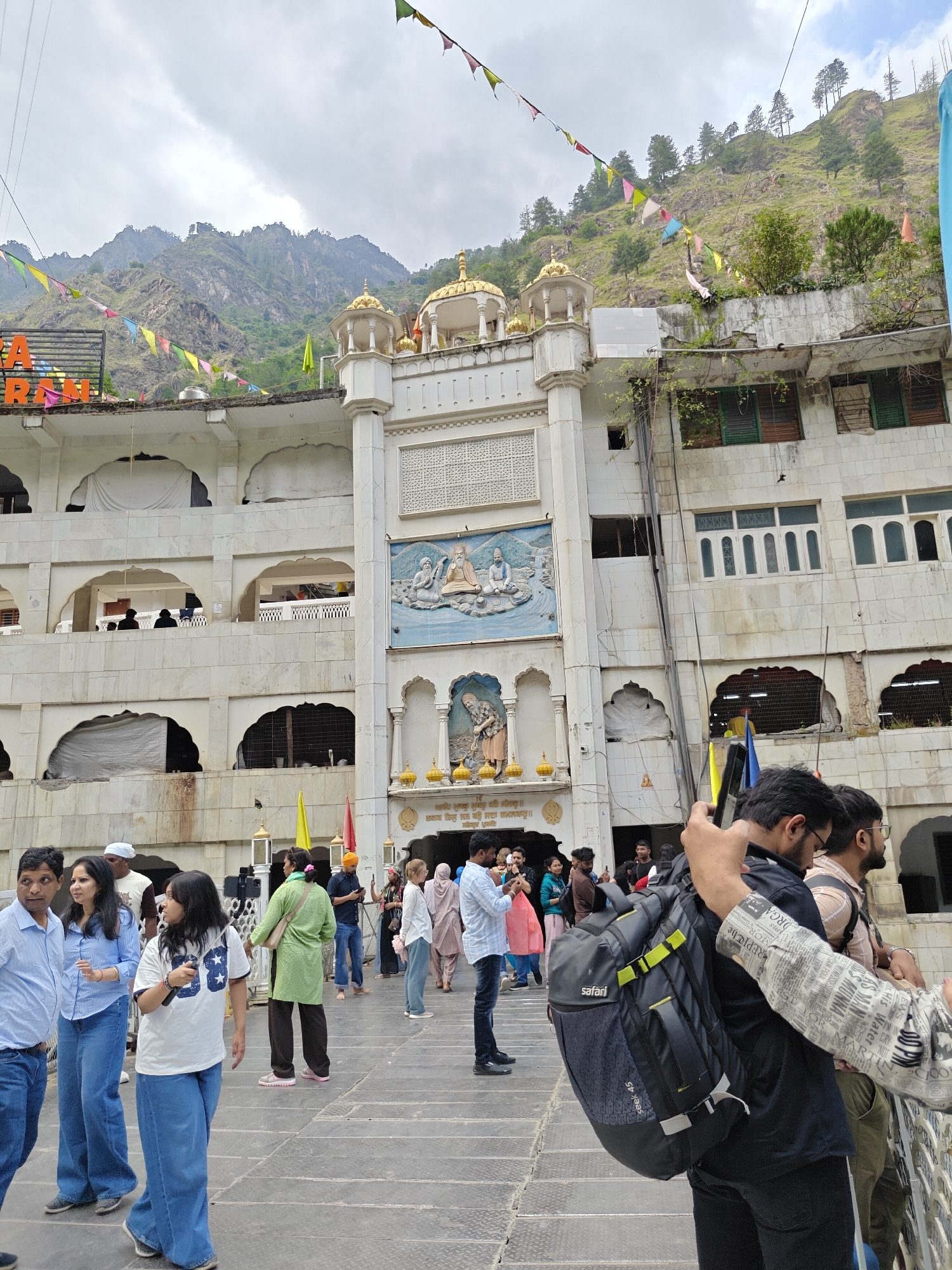 This outdoor photograph captures a bustling scene at Gurudwara Shri Manikaran Sahib in Kasol, India. The central subject is a large, multi-story Gurudwara building, characterized by its white facade, traditional Sikh architectural elements, and golden domes adorning its roof. The main entrance features elaborate relief sculptures depicting religious figures, with arched windows and openings visible on multiple levels. The Gurudwara is dramatically set against a backdrop of a steep, green, rocky mountain, sparsely covered with trees, highlighting its serene and natural location.
Numerous colorful prayer flags, or pennants, are strung across the scene, draped both in front of the Gurudwara structure and extending upwards into the mountain's greenery, contributing to a spiritual and festive atmosphere. The paved courtyard in the foreground is filled with a diverse crowd of people. Casually dressed individuals of various ages are seen walking, standing, and congregating, indicating a busy day of visitation or pilgrimage at this sacred site.
In the immediate foreground, a man and two women are walking on the left, while on the right, a man with a prominent black "Safari" brand backpack is visible. The sky is overcast, suggesting cloudy daytime weather, yet there is sufficient natural light. A significant detail is the inscription in Punjabi script on the building: "ਸਲਕ ਤਿਸੁ ਜਨ ਕਉ ਸਦਾ ਨਮਸਕਾਰੁ।" (Salok Tis Jan Kau Sada Namaskaar), a Sikh scripture verse translating to "Salutations always to that person/devotee." Yellow and red flags are also visible near the building's entrance. The image conveys the vibrant atmosphere of a popular religious landmark nestled within a mountainous region.