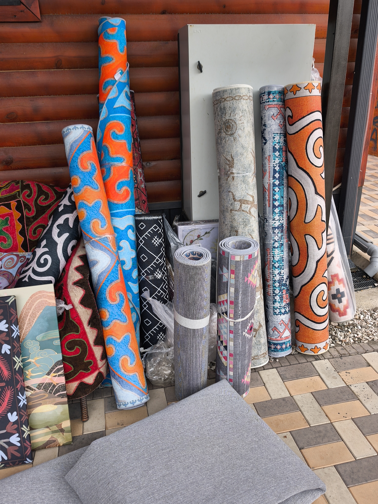 This image depicts an outdoor display of numerous rolled rugs and carpets, likely at a market stall or shop entrance in Karakol City, Kyrgyzstan. The rugs exhibit a wide variety of colors, patterns, and sizes, creating a vibrant and textured scene.
The most striking rugs feature bold, curvilinear patterns in bright blue and orange, often outlined in white. These are seen in several large, vertically rolled pieces. Other notable designs include a black rug with intricate white geometric patterns, and several lighter-colored rugs with more subtle motifs, such as a grey one with repeated deer or antelope figures. Smaller rolled rugs display patterns in shades of grey, pink, and black, some with abstract or tribal-like designs. A few partially unrolled pieces reveal colorful patterns in red, black, and beige.
The rugs are leaning against a rustic wooden plank wall, which forms the primary background for the display. To the right, a white electrical box with the text "EKT" is mounted on the wooden wall. The ground beneath the display is a mosaic of multi-toned paving stones in rectangular shapes, colored in varying shades of brown, beige, and grey. Further to the right, the paving transitions to an area of gravel and a visible drainage pipe. In the immediate foreground, two folded pieces of grey fabric or cushions are resting on the paving stones.
The scene appears to be well-lit by natural daylight, with no harsh shadows, suggesting good weather conditions. No people are visible in the image, implying the activity is primarily the presentation or storage of these textile goods. The overall impression is one of a colorful and inviting collection of artisanal or commercially produced floor coverings.