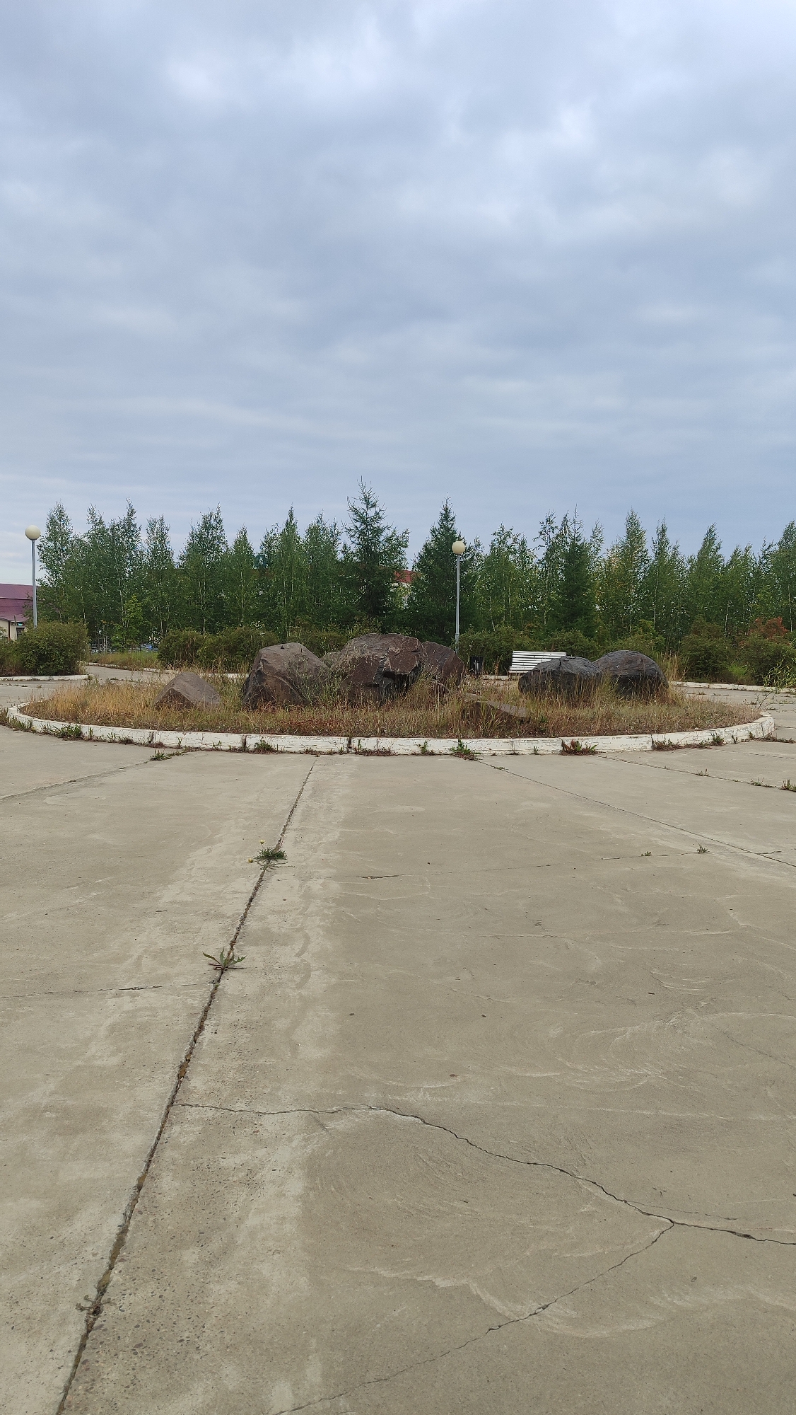 This is an outdoor, eye-level, full shot of a concrete area with a circular flower bed in the center, located in Mirny, Russia. The flower bed contains several large, dark rocks and dry grass. In the background, a line of deciduous trees and some evergreen trees forms a backdrop. Two lampposts are visible within the scene, one on the left and one on the right. A white bench is situated behind the flower bed. The sky above is overcast with gray clouds. The concrete surface in the foreground shows cracks and a single plant growing from one of the cracks.