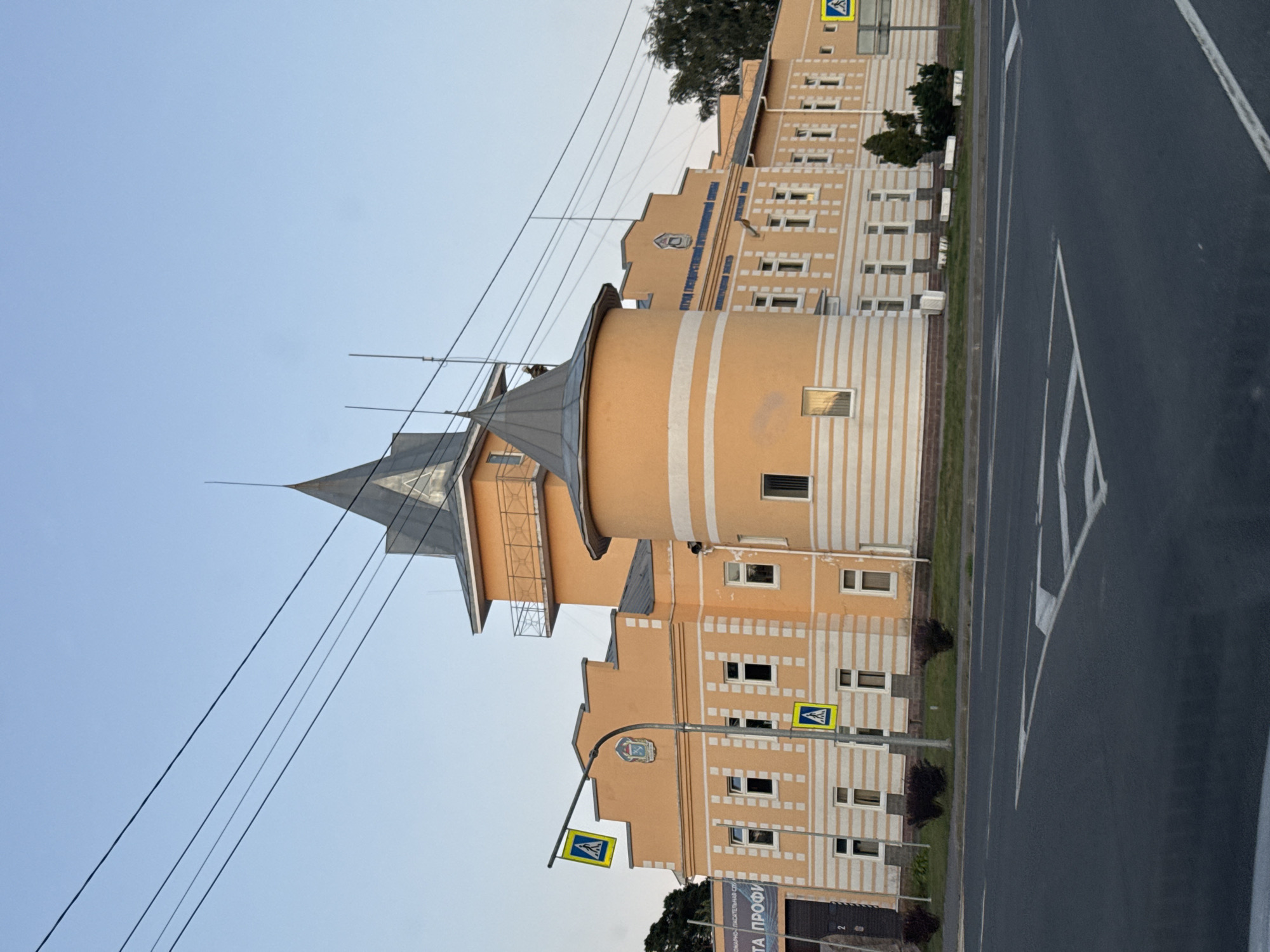 The image shows a two-story, orange-colored building with a distinctive architectural style, featuring a rounded tower and a conical roof, reminiscent of a castle. The building is located in Priozersk, Russia, and appears to be a public institution, as suggested by signage. A pedestrian crossing sign is visible in front of the building, indicating its proximity to a road. The sky is clear and blue, and power lines are strung across the upper portion of the frame. The road in the foreground has visible lane markings. The overall impression is one of a calm, daytime scene in a semi-urban environment. There are no people visible in the image.