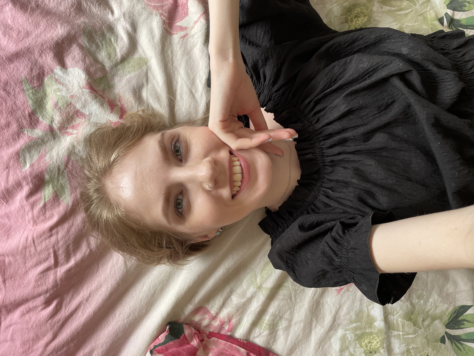A young woman is captured in a close-up, overhead shot, lying on a bed in what appears to be an indoor setting. She has fair skin, light blonde hair, and striking light-colored eyes, and she is smiling broadly at the camera, revealing her teeth. She wears a black top characterized by a ruffled or smocked neckline and similarly detailed cuffs. Her left hand is raised, with fingers gently touching her chin and cheek, adding a relaxed and playful element to her pose.
The bedding beneath her features a distinct floral pattern, predominantly in shades of pink, white, and light green, suggesting roses or similar blooms. Visible accessories include a silver-toned hoop nose ring and a delicate silver chain necklace. Small earrings can also be seen in her left ear. Her fingernails are visible, appearing to be well-maintained and possibly painted with a light or clear polish.
The overall mood of the image is cheerful and relaxed, conveyed by her radiant smile and casual pose. The lighting is bright and soft, indicating an indoor daytime environment. There is no visible text within the image. This scene is set in Izhevsk, Russia.