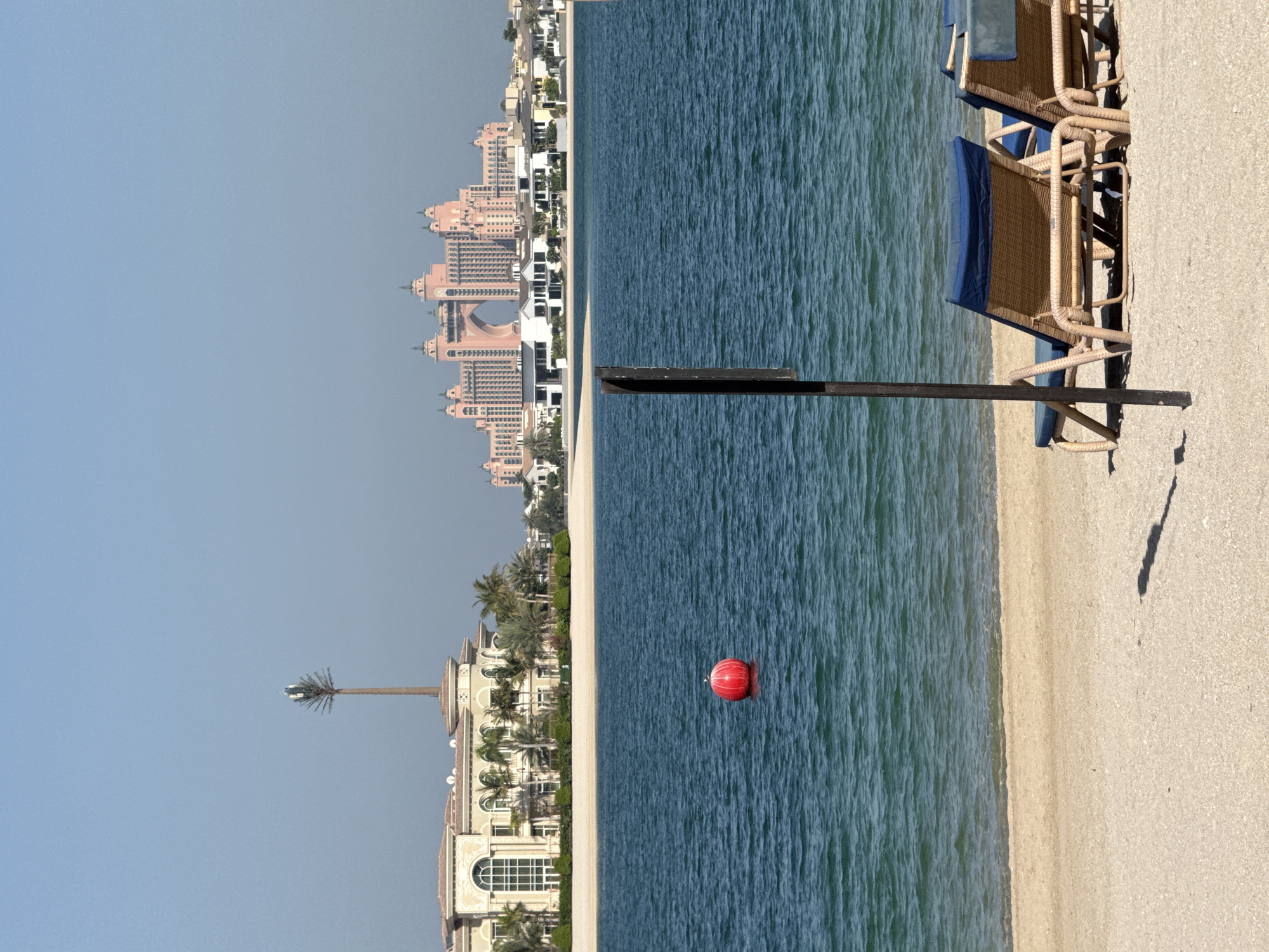 This is a photograph taken from a beach in Dubai, United Arab Emirates, looking out over the water towards a cityscape. In the foreground, two empty lounge chairs with blue cushions are positioned on a sandy beach, with a dark, thin pole casting a shadow to their left. The ocean is a deep blue, with gentle waves. A bright red buoy is visible in the water. In the background, a large, pink resort complex with multiple towers and an arched central structure dominates the skyline, characteristic of the Atlantis, The Palm hotel. To the left of the resort, another building with a beige facade and large arched windows is visible, possibly part of the same resort or a nearby hotel. A tall, slender structure resembling a palm tree with visible antennas at its top stands near the beige building, likely a disguised cellular tower. The sky is clear and blue.