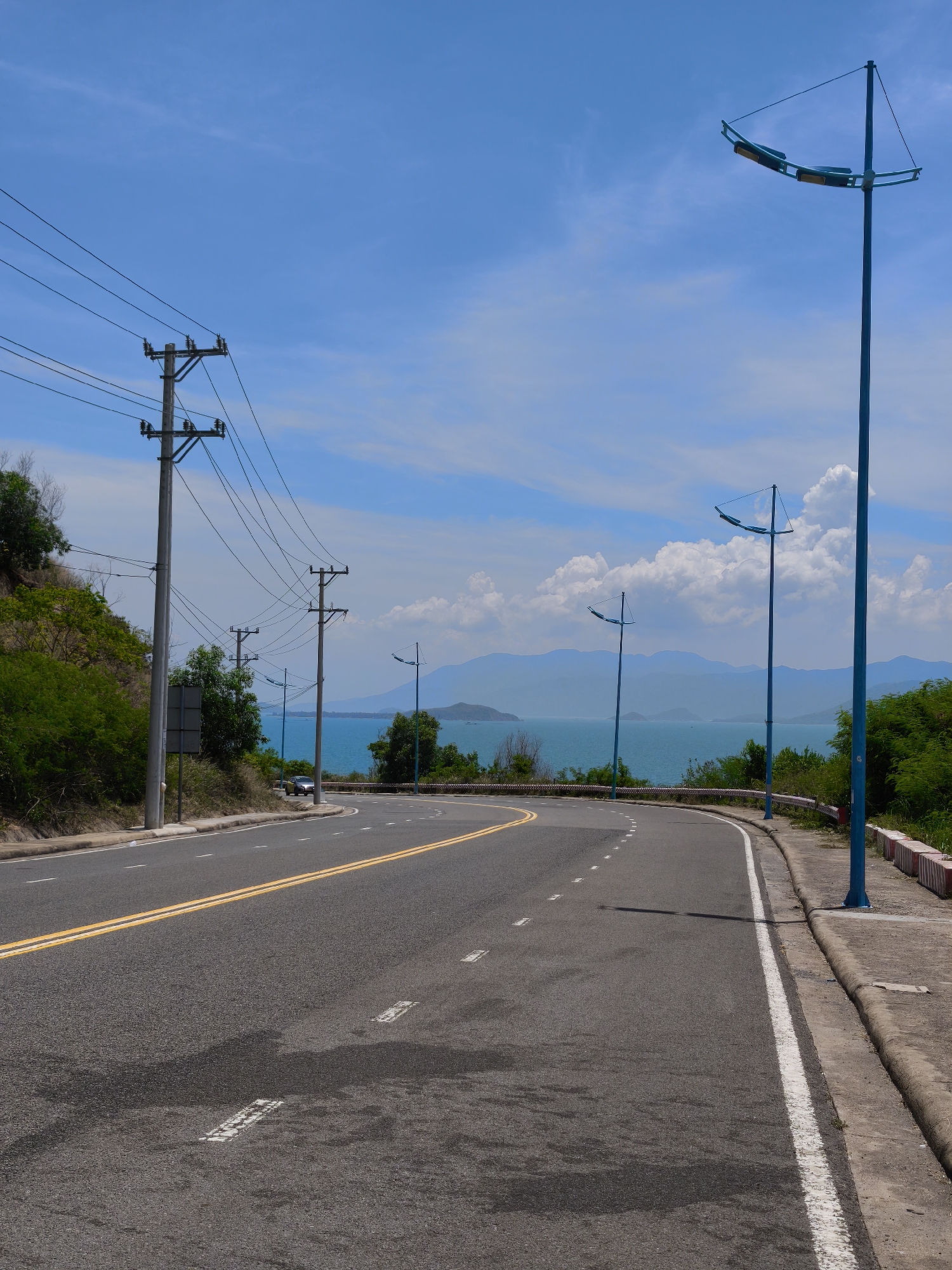 The image shows a road curving along a coastline in Nha Trang, Vietnam. The road is asphalt with yellow double lines and white dashed markings. On the left side of the road, there is a steep, vegetated hill with utility poles and power lines. On the right side, there is a concrete curb and sidewalk, with three blue streetlights spaced along the road. In the distance, the turquoise ocean stretches to the horizon, with hazy mountains visible across the water. The sky is blue with scattered white clouds. The scene is peaceful and scenic, with no people or activity visible in the immediate vicinity, though a car can be seen in the distance on the road.