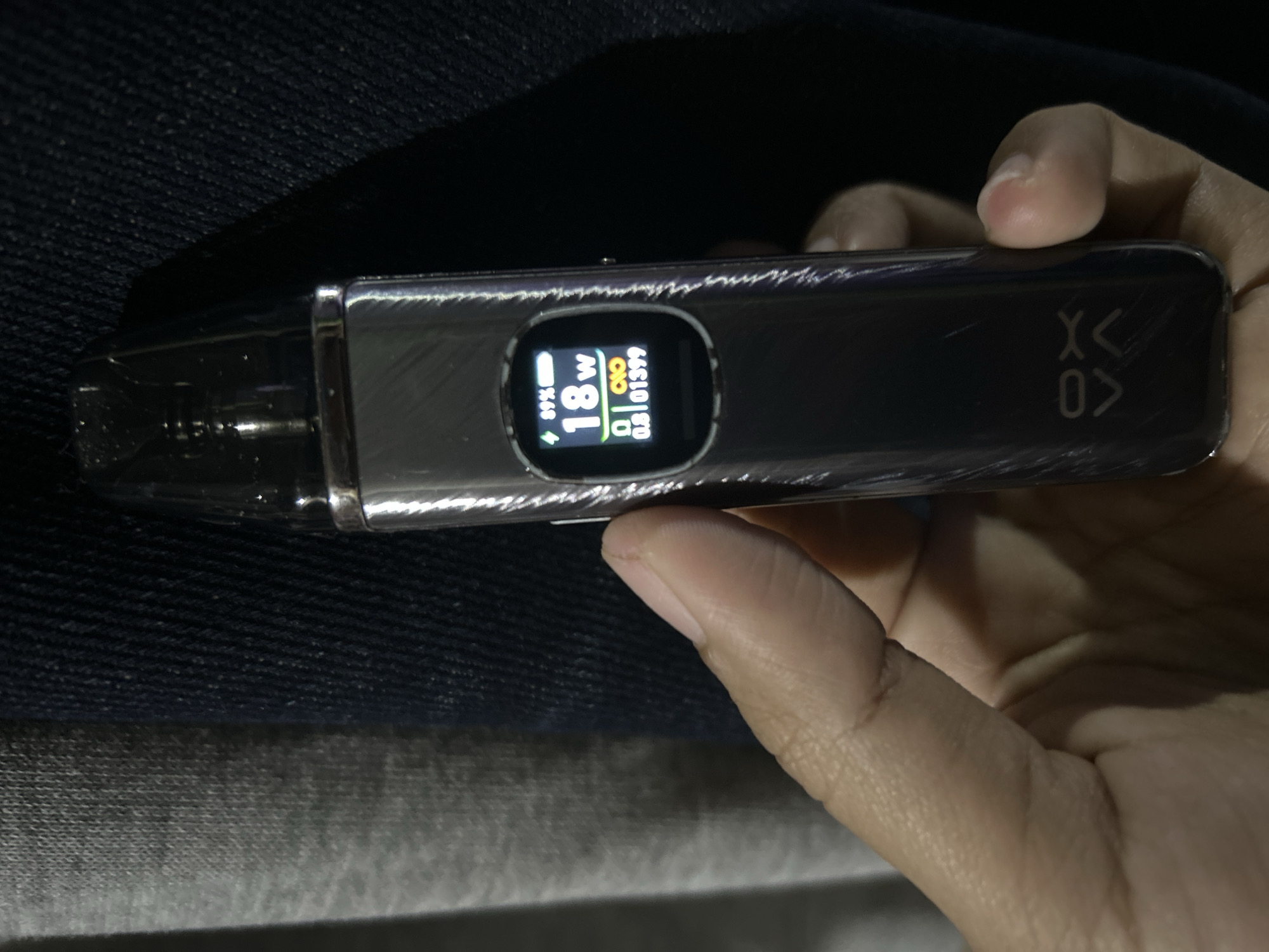 The image shows a hand holding a metallic vape pen with a digital display. The display shows "89%" for battery, "18W" for wattage, and "0.8 Ω 01399" for resistance and puff count. The vape pen has a logo with "XV" on its side. The background consists of dark blue denim fabric and a grey textured fabric. The lighting suggests an indoor or dimly lit outdoor environment. The image is a close-up shot, focusing on the vape pen and the hand. There is no identifiable location in Depok, Indonesia.
