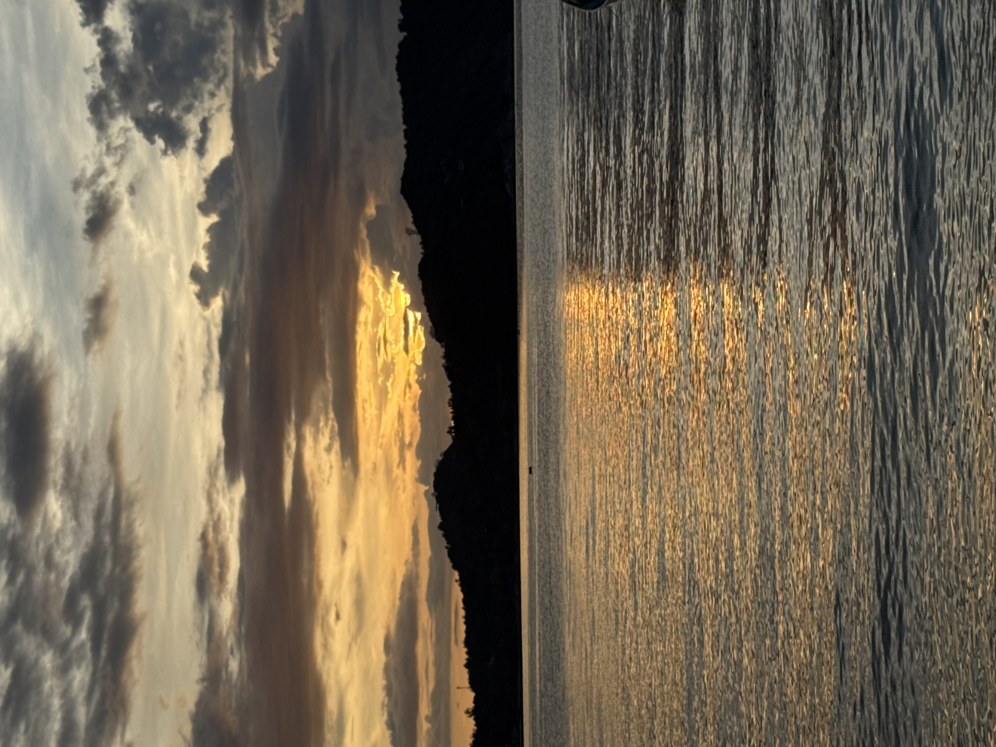 The image depicts a sunset over the water in Koh Phangan, Thailand. The sky is filled with dramatic clouds, with the sun peeking through a gap, casting a golden glow. A dark, silhouetted landmass, likely a hill or island, forms the horizon line. The water in the foreground is calm, reflecting the colors of the sunset. A small, dark object, possibly a boat or buoy, is visible in the distance on the water. Part of a boat is visible on the far right edge of the frame.