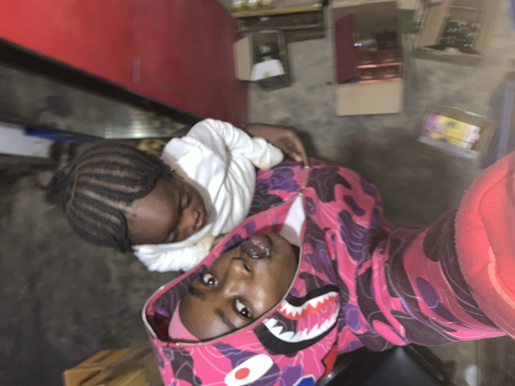 The image appears to be a selfie taken indoors. A person wearing a pink and purple camouflage hoodie is holding the camera, with a younger child resting on their chest. The child is wearing a white shirt and has braided hair. The background shows boxes and what appears to be a red refrigerator or display case. It's difficult to determine the exact location within New Stadium, Nigeria, without more context.