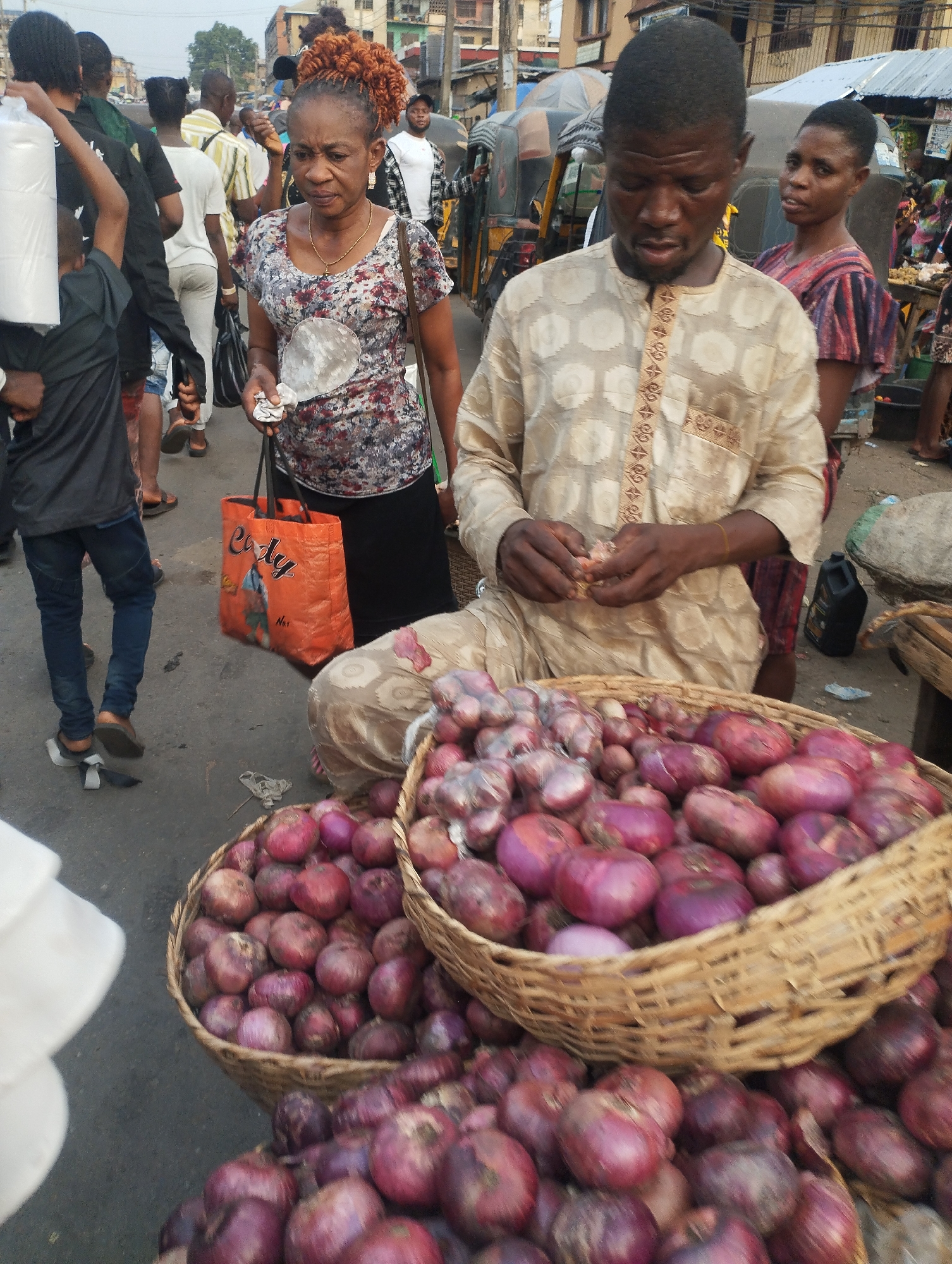 The image depicts a busy street scene in Fegge, Nigeria. In the foreground, a man is seated beside baskets overflowing with red onions. He appears to be handling or sorting the onions. An older woman wearing a floral top and black skirt is standing nearby, holding a clear plastic object and an orange shopping bag with the word "Curly" on it. Behind her, there are several other people walking through the scene, including a child carrying rolls of white paper. A woman in a patterned red dress is also standing close to the onion seller. In the background, there are buildings, motorbikes, and other vehicles suggesting a crowded marketplace or commercial area. The street is paved but appears somewhat worn.