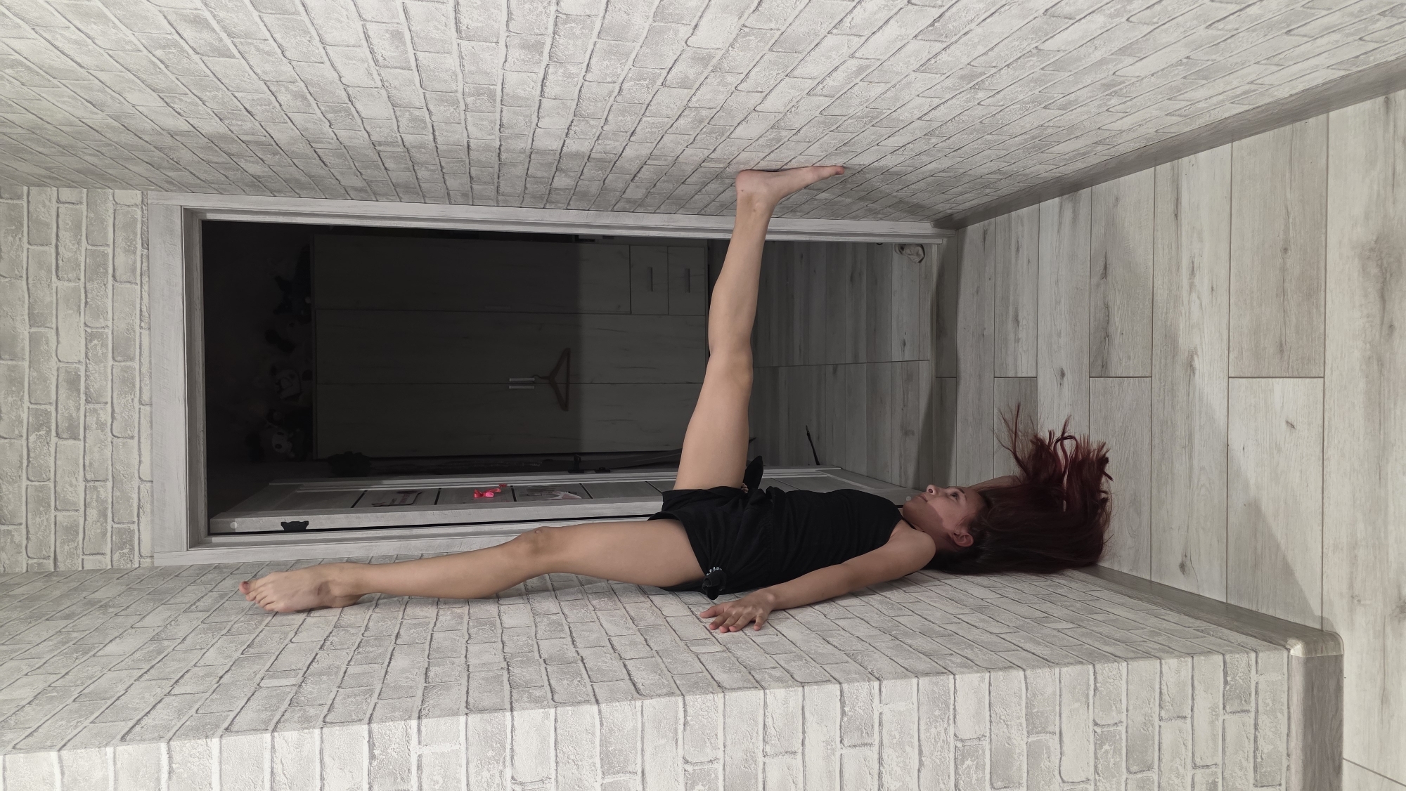 A woman lies on a surface that resembles a brick wall, which may actually be flooring. She wears a black top and dark shorts. One leg is extended upward, nearly perpendicular to the surface, while the other leg is outstretched along the floor. Her head rests near what appears to be a wooden wall or wainscoting. The background shows a doorway leading to a dark interior space. The ceiling also appears to be covered in brick-like material, suggesting an unconventional orientation of the scene. The location is in the city of Unknown, Unknown.