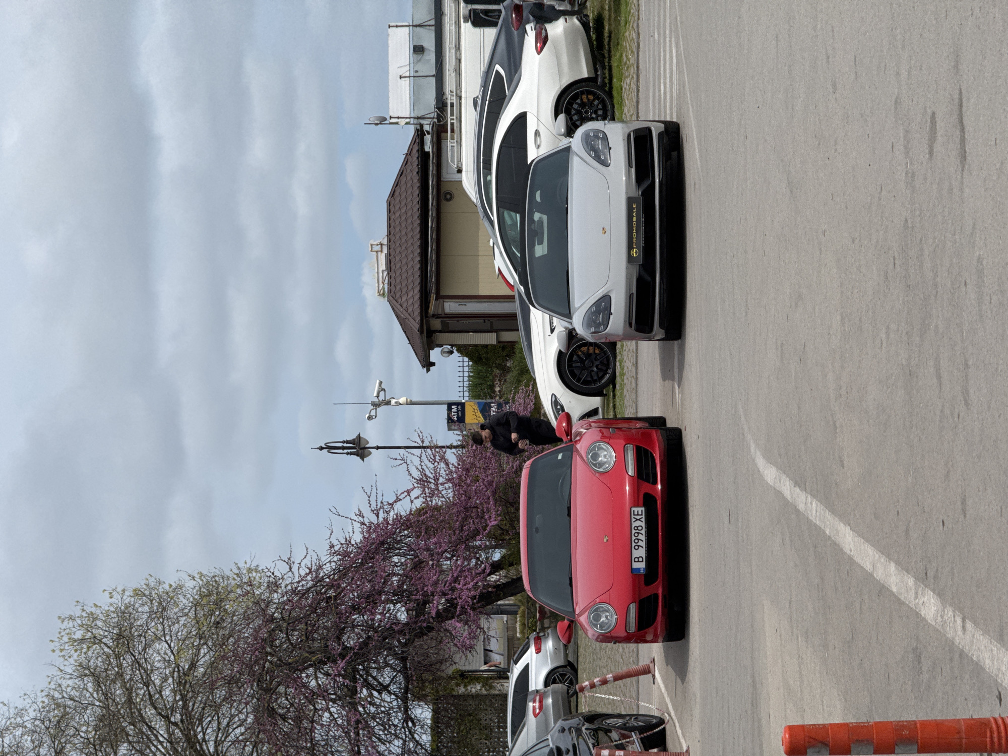 The image depicts an outdoor parking area on an overcast day, likely in Varna, Bulgaria, given the license plate and architectural cues. Several cars are parked on asphalt marked with white lines.
In the foreground, a vibrant red Porsche 911 (997 generation) is prominently featured, facing away from the viewer. Its Bulgarian license plate reads "B 9998 XE". Behind it, a white Porsche Panamera (971 generation) is parked, displaying a "PROMOSALE" banner on its front bumper. Further back, several other white and dark-colored cars are visible.
A person, dressed in dark clothing, is seen bent over near the front passenger side of the red Porsche, appearing to be inspecting something or interacting with the car.
To the left of the cars, a large tree with distinctive pink/purple blossoms adds a touch of color to the scene, suggesting springtime. Another tree with lighter green leaves is also visible. In the midground, a low-rise building with a brown tiled roof and light-colored walls stands, behind which more parked vehicles can be seen.
Above the buildings and trees, the sky is largely overcast with soft, diffuse light, indicating daytime. Visible infrastructure includes a streetlight pole with multiple security cameras attached, and a sign displaying "ATM" along with Cyrillic text below it. Orange traffic cones or barriers are present on the right side of the frame, bordering the parking lot.