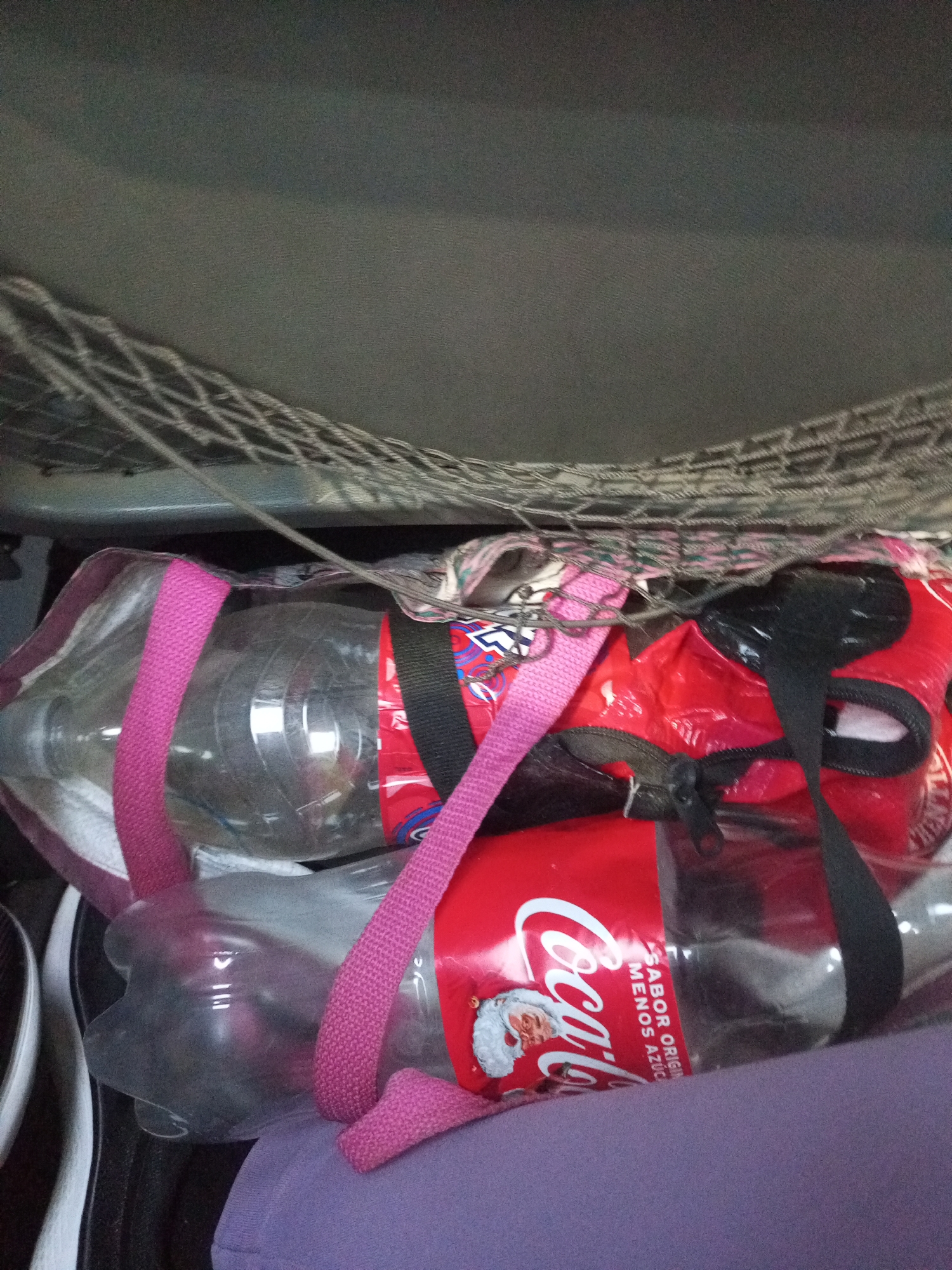 Two clear plastic bottles are contained within a patterned tote bag. One bottle, on the left, is partially filled with a clear liquid and has a pink strap across it. The second bottle, on the right, contains a clear liquid and is adorned with a red label featuring the Coca-Cola logo and an image of Santa Claus. The bag also has a black strap and a zipper. The items are situated inside a vehicle, likely in the trunk, as indicated by the presence of a cargo net above. The surrounding environment appears to be dark and confined. The image was taken in Juan Griego, Venezuela.