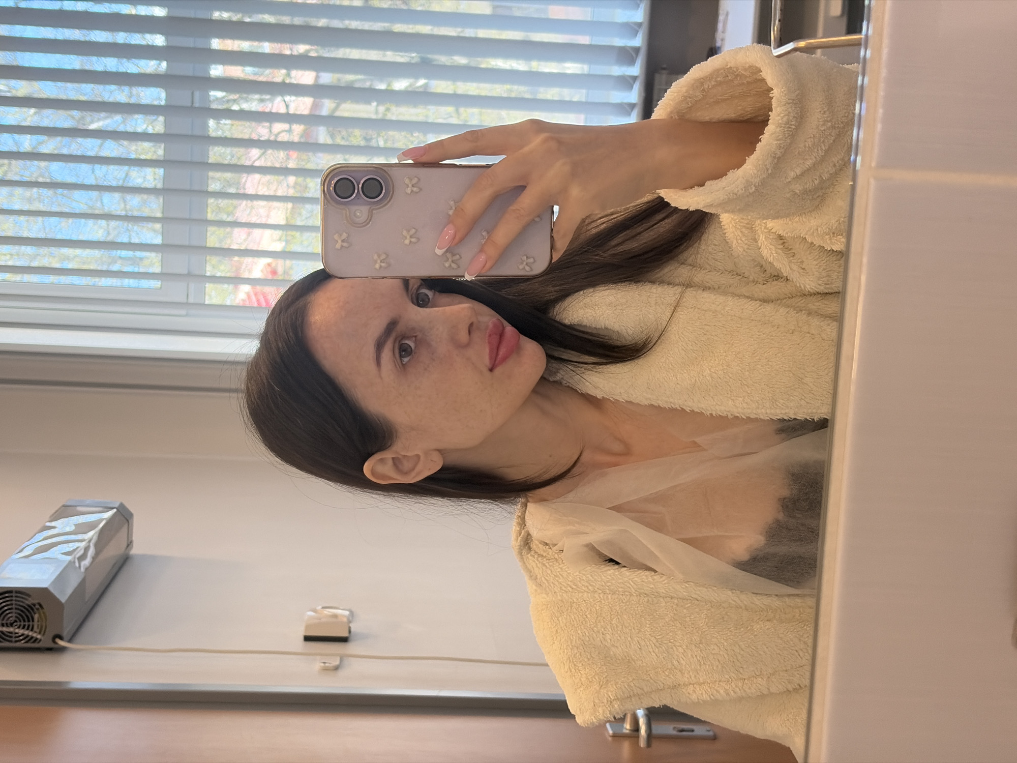 A woman is taking a selfie in front of a mirror. She is wearing a beige bathrobe. The woman is holding a cellphone with a light purple floral case. In the background, there is a window with white blinds. Outside the window, trees and a building are visible. A hair dryer is affixed to the wall. The location is Saint Petersburg, Russia.