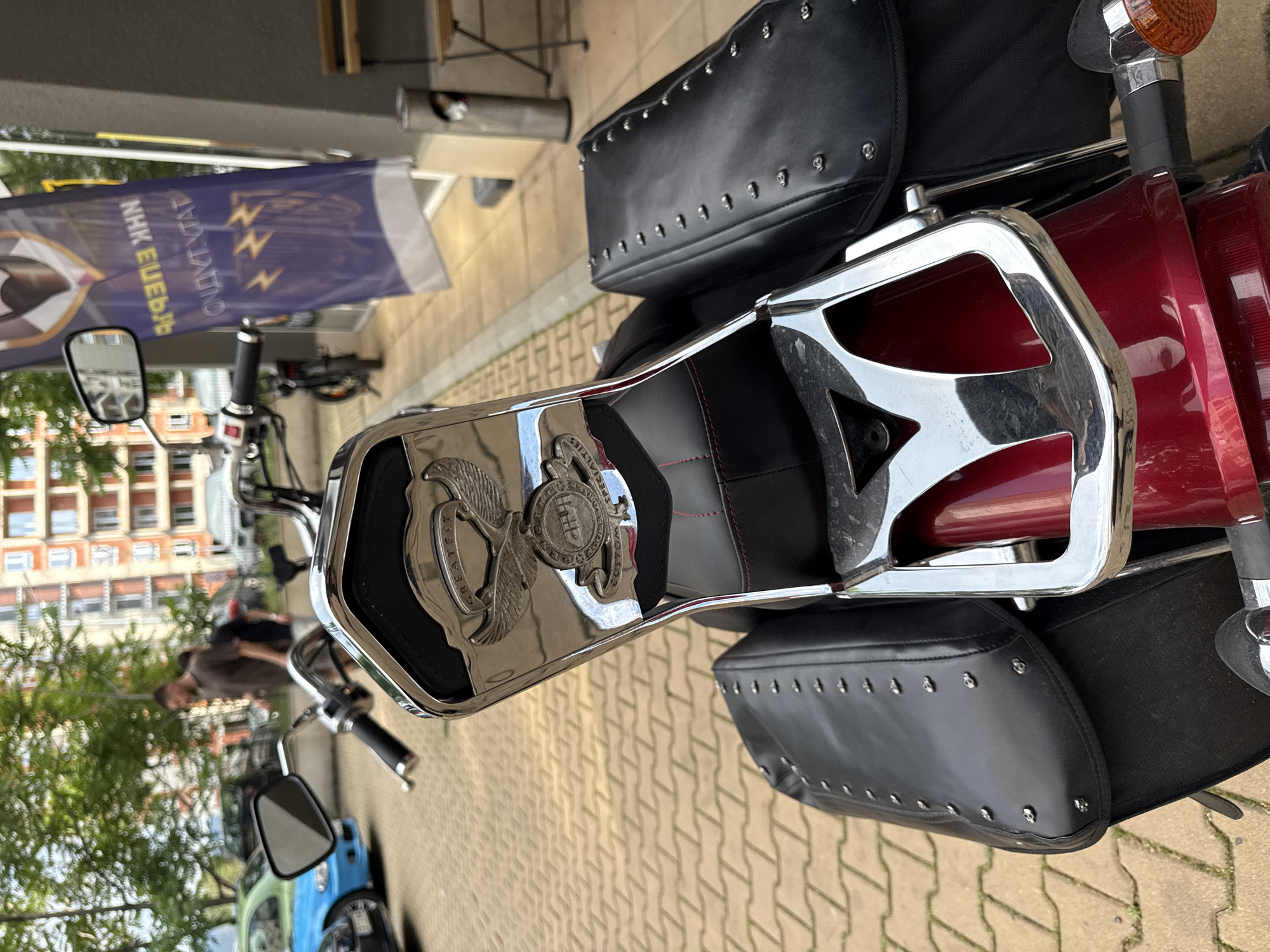 A deep red motorcycle is parked outdoors, viewed from the rear, on light-colored interlocking pavers. The motorcycle features extensive chrome, including a backrest with a padded black seat detailed with red stitching. A prominent chrome emblem on the backrest displays an eagle with outstretched wings, flanked by the word "GREATEST" above and a circular crest with partially visible text like "CUSTOM SPECIAL" below. Black, studded leather-like saddlebags are mounted on either side of the rear fender. The right rear turn signal, with an orange lens, is visible at the bottom right. In the blurred background to the left, a light blue car with a partial green paint job is parked, alongside other vehicles. Two men are walking in the mid-ground, one in a dark t-shirt and another further back in a white t-shirt. Behind them is a multi-story building with a light-colored facade and numerous windows. Green foliage from trees is visible at the top left. To the right, a dark gray building facade is visible, with a vertical purple/blue flag hanging. The flag displays mirrored Cyrillic text, appearing as "NHH ЕЛЕКТРО" and "ЕЛЕКТРОНИКА" (Electronics), along with three lightning bolt symbols, indicating a commercial establishment in Sofia, Bulgaria. A stainless steel trash receptacle and a foldable outdoor chair are visible near the flag.