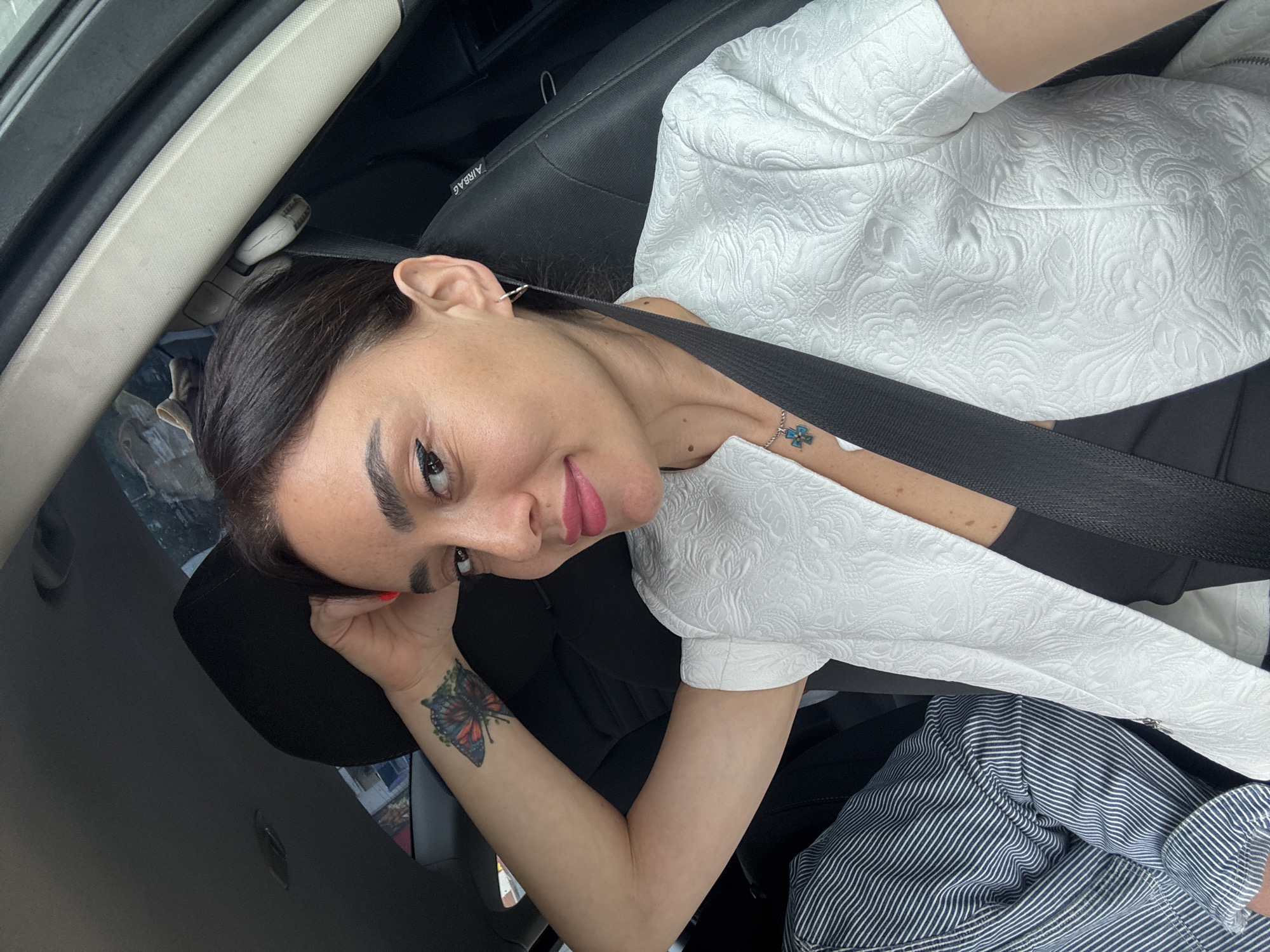 A woman with dark hair, winged eyeliner, and pink lipstick is pictured inside a car. She is wearing a white textured jacket over a black top and a silver necklace with a turquoise cross pendant. A seatbelt is visible across her chest. Her left arm is bent, resting her hand on the back of the car seat, revealing a butterfly tattoo on her forearm. She is looking towards the camera with a slight smile. The background shows the interior of a car with black upholstery and a glimpse of the outside environment through the windows.