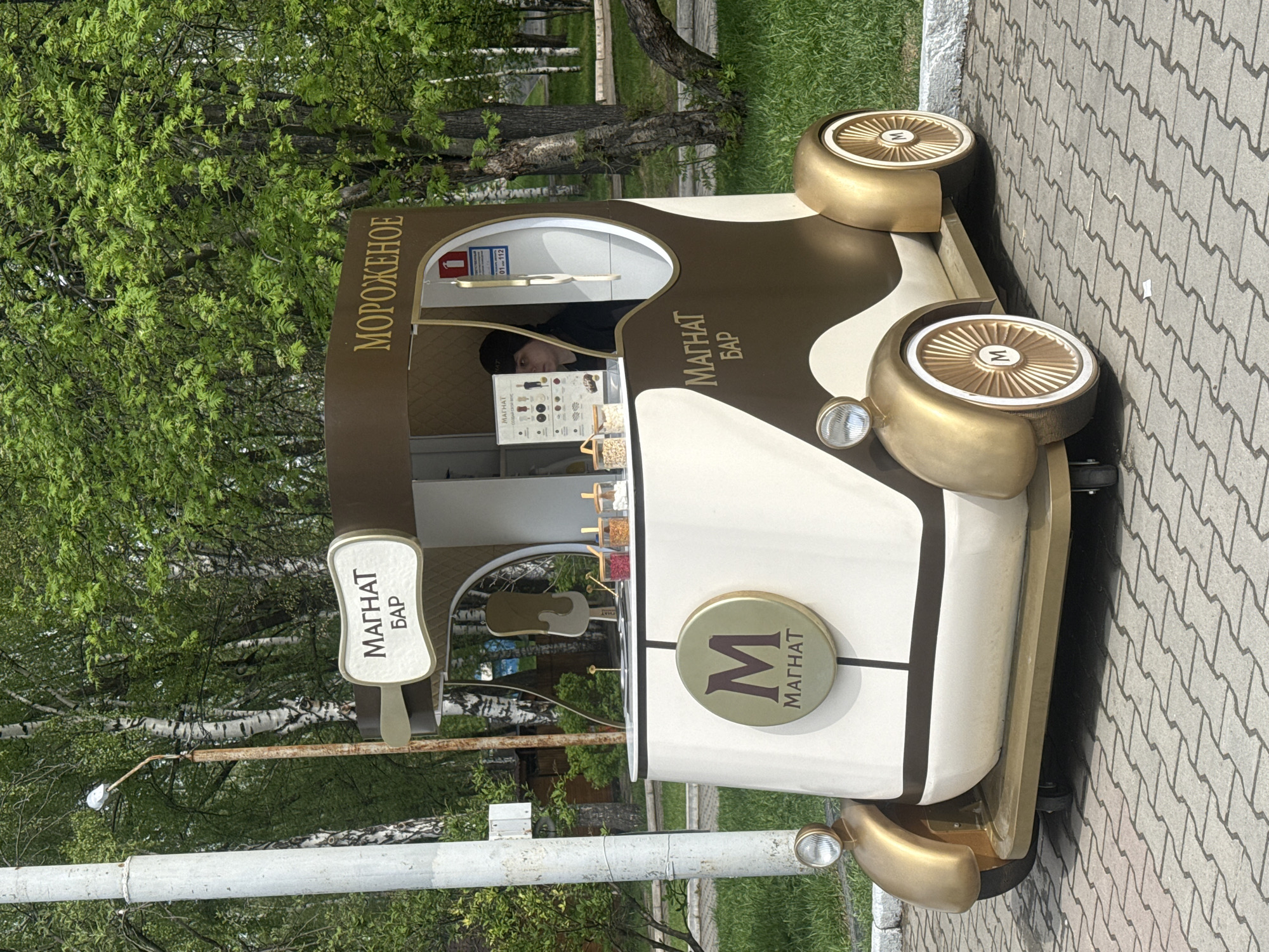 A close-up, rotated, ground-level shot depicts a decorative ice cream vendor's cart in Yekaterinburg, Russia. The cart, adorned in gold and white with brown accents, features "МОРOЖEHOE" in Cyrillic script and "МАГНАТ БАР" printed on its sides. The wheels are notably large and gold-colored. A vendor wearing a black hat stands inside the cart, behind a counter showcasing various ice cream toppings. The surrounding environment includes trees, foliage, and a paved walkway. A utility pole is visible in the background.