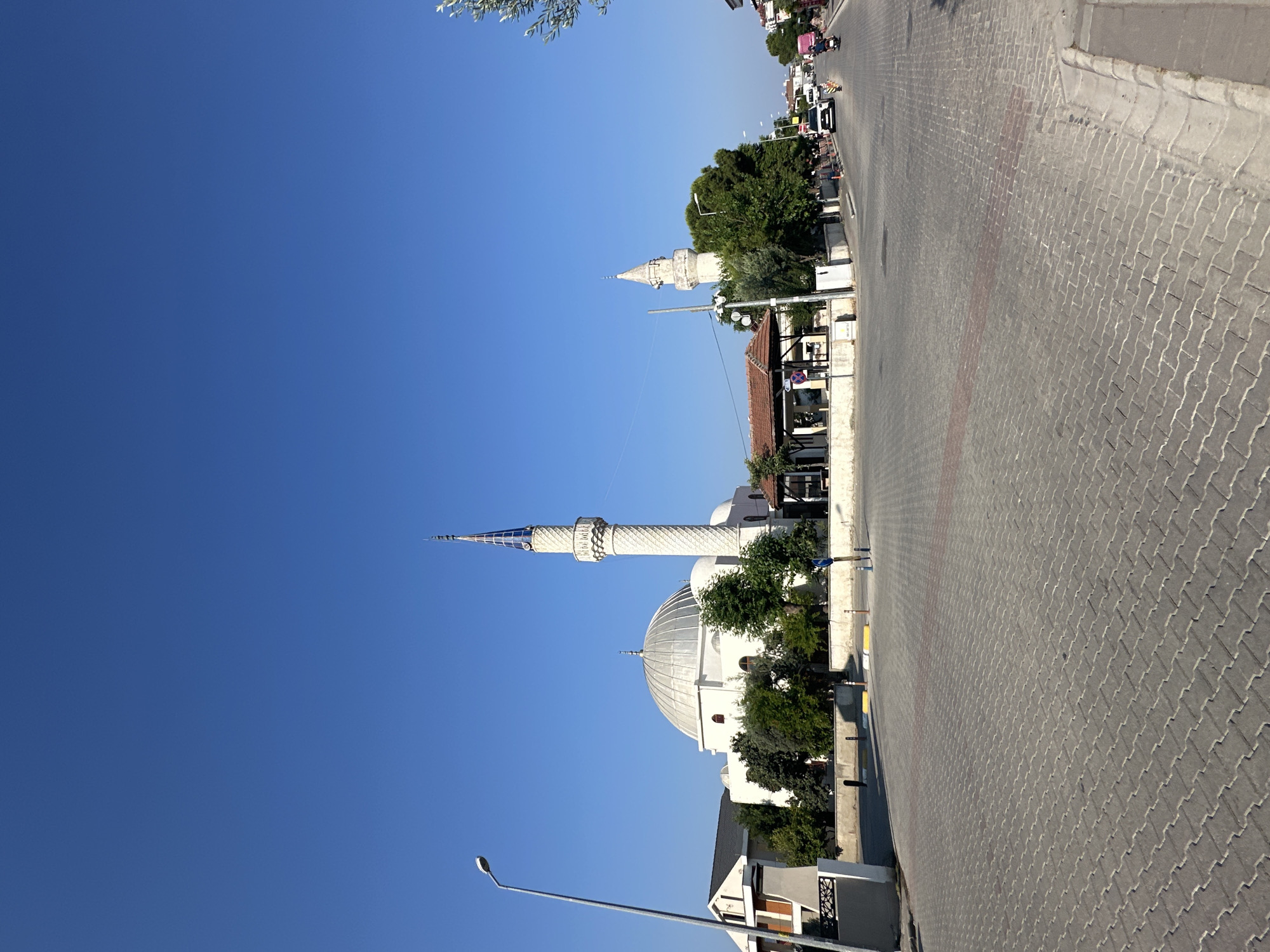 The image depicts a street scene in Fethiye, Turkey, featuring a prominent mosque with a large dome and a tall minaret. The mosque is white and situated among trees. To the left of the mosque, a modern building with a slanted roof is visible. In the foreground, a paved street with interlocking bricks runs diagonally across the frame. The street is lined with trees and a sidewalk. Several vehicles, including cars, a motorcycle, and a pink scooter, are on the street, suggesting moderate traffic. The sky is a clear, bright blue, indicating a sunny day.