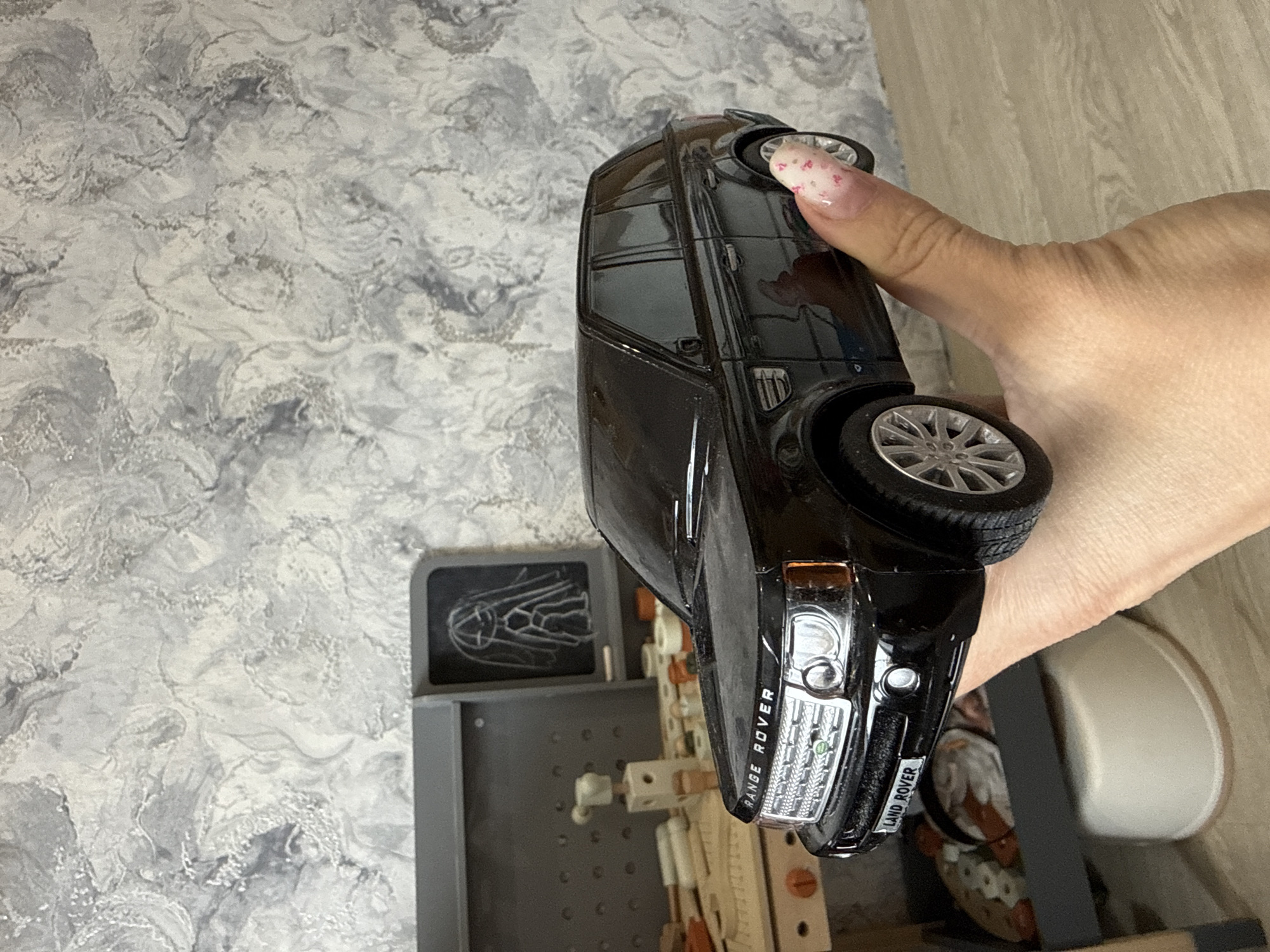 The image shows a black toy Land Rover Range Rover held in a person's right hand. The background includes a grey textured wall on the left and a wooden surface on the right, with a grey pegboard and some miscellaneous small items visible below the toy car. The location is not evident in the image, and there is no information linking it to Rusanovo, Russia.