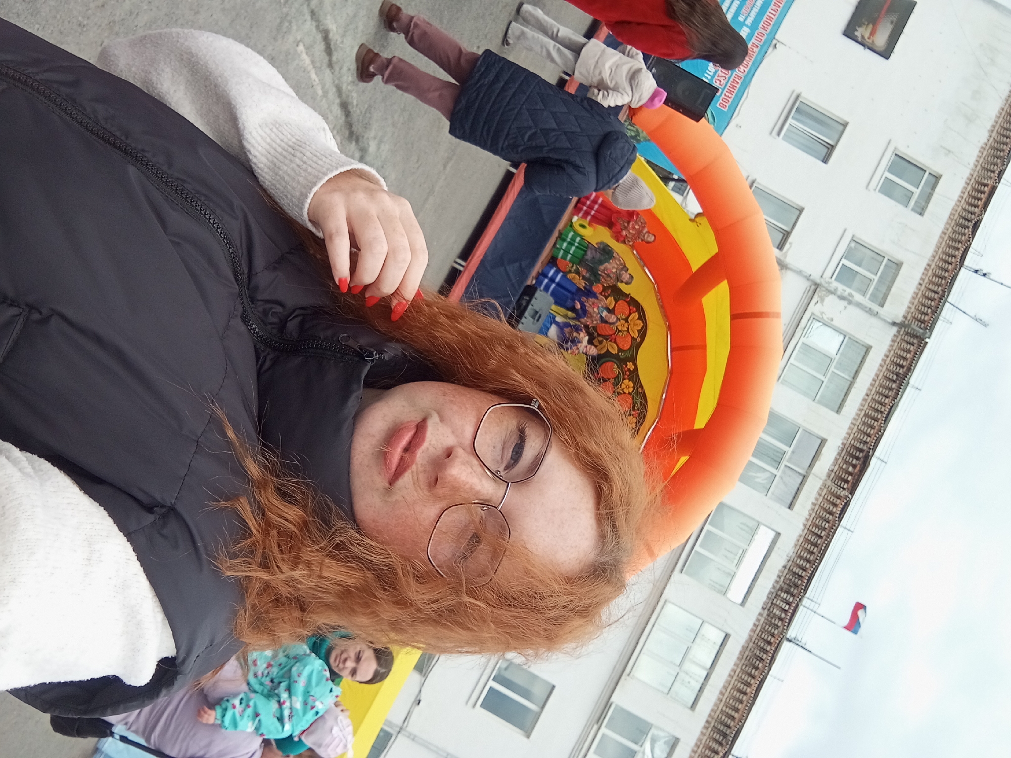 The image presents a person with red hair and freckles, wearing glasses and a black puffy vest over a white sweater. The person has red nail polish. The background includes an inflatable orange archway with colorful painted details, a building with multiple windows, and a Russian flag. There are other people visible, including some partially obscured in the lower left corner and some standing near the inflatable arch. The image appears to be taken in Yurgamysh, Russia.