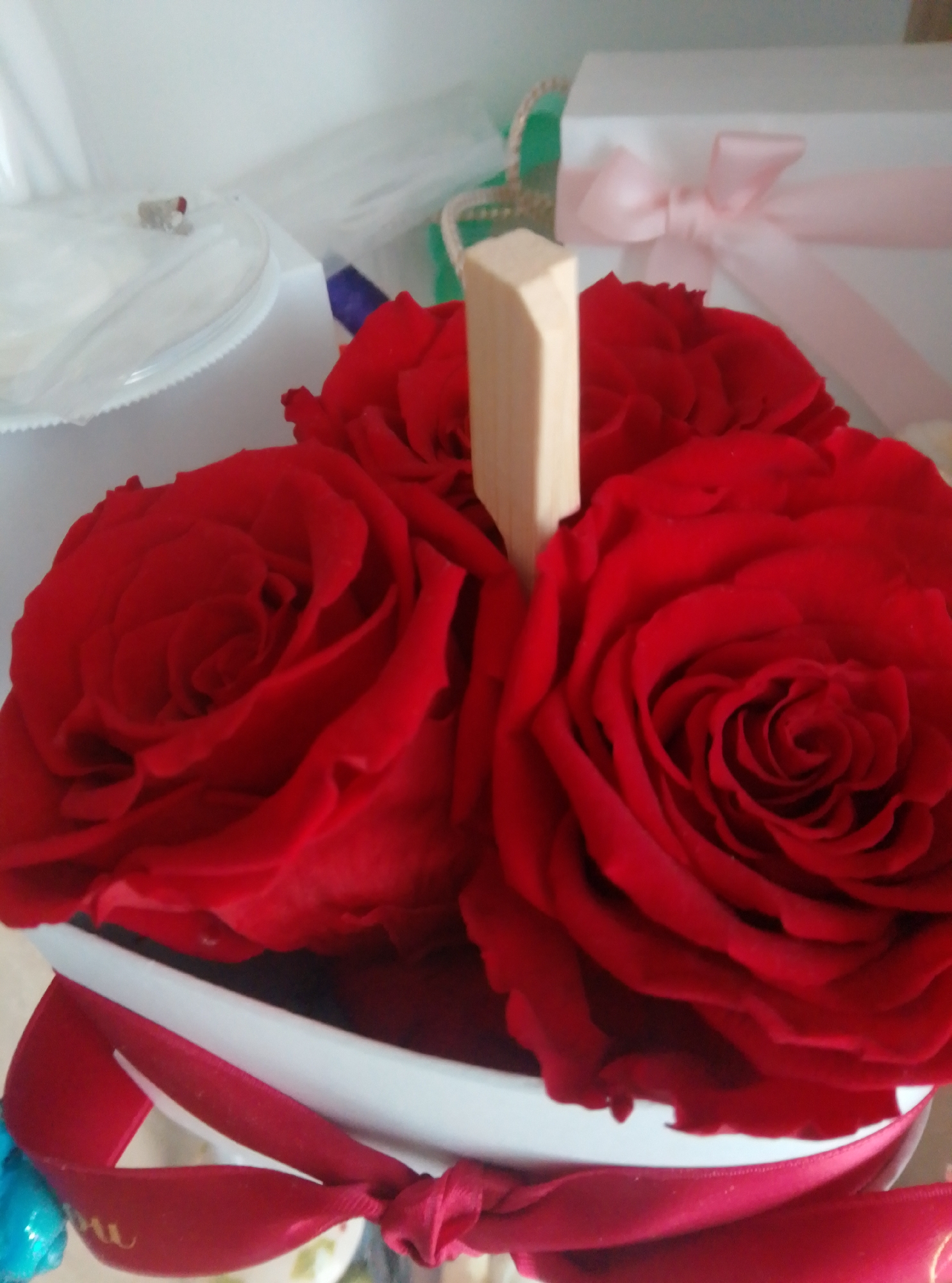 A close-up, overhead shot features a floral arrangement of three large, deep red roses. The roses are arranged in a white bowl or container, which is partially obscured by red ribbon tied around its rim. A small, light brown wooden stake protrudes from the center of the roses. In the blurred background, a white gift box with a pink ribbon is visible, along with other indistinct objects. The setting appears to be indoors, possibly a room in a residential or commercial space in the city of Wedel, Germany. The lighting is soft and diffused.