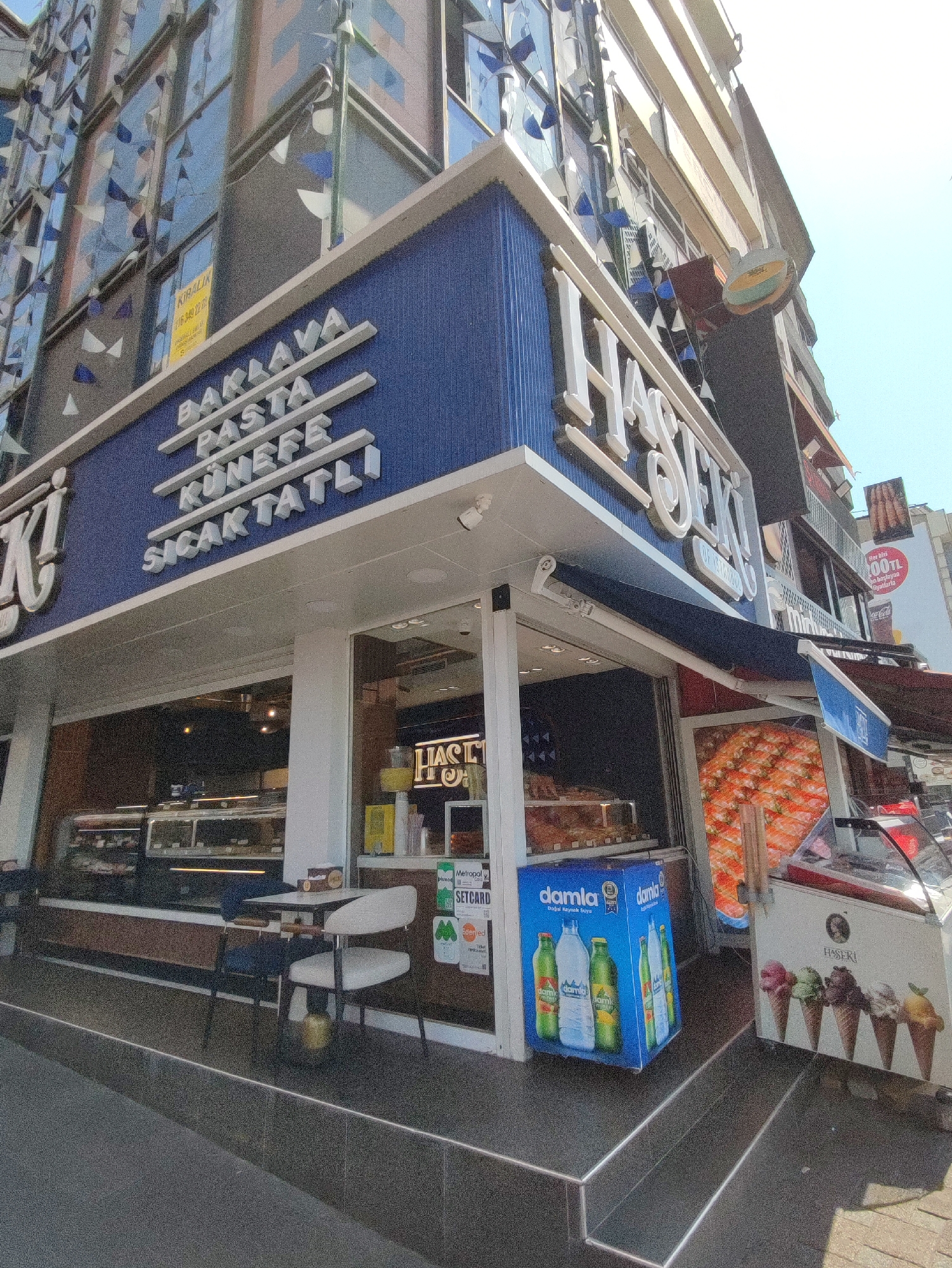 This is a daytime, low-angle, outdoor, eye-level shot of the exterior of a dessert shop in Kadıköy, Turkey. The shop, named "Haseki", is visible with its blue and white facade adorned with white, 3D lettering that reads "BAKLAVA PASTA KÜNEFE SICAK TATLI" on the left and "HASEKİ" on the right. There are windows displaying various desserts and ice cream. In front of the shop, there is an ice cream freezer displaying pictures of ice cream cones and a blue cooler box with bottles of water. Several tables and chairs are set up outside for customers. The street in front of the shop is paved with dark stone. In the background, there is a multi-story building with windows decorated with triangular flags. A yellow "KİRALIK" (for rent) sign is visible on one of the windows of the building. Another sign in the background advertises a discount on "200TL" purchases. The sky is clear and blue. There are no people clearly visible in the frame.