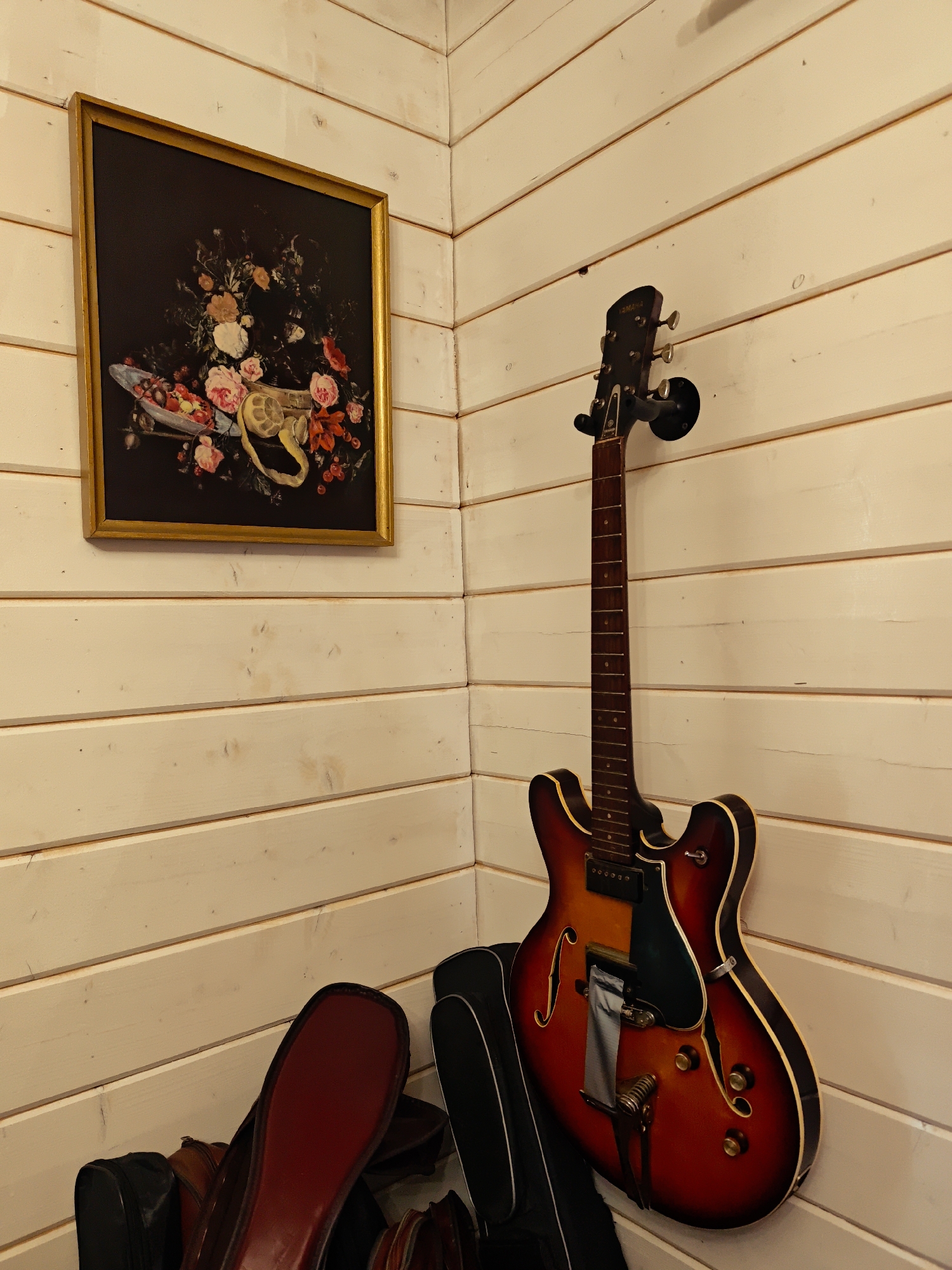 An interior corner of a room in Свистуха (кузяевский С/о), Russia features horizontal, white-painted wooden plank walls. On the left wall hangs a framed still-life painting with a gold frame depicting flowers and fruit on a dark background. On the right wall, an electric guitar hangs from a black wall mount, its body leaning against several instrument cases on the floor. The guitar is red and brown sunburst with a dark fretboard.