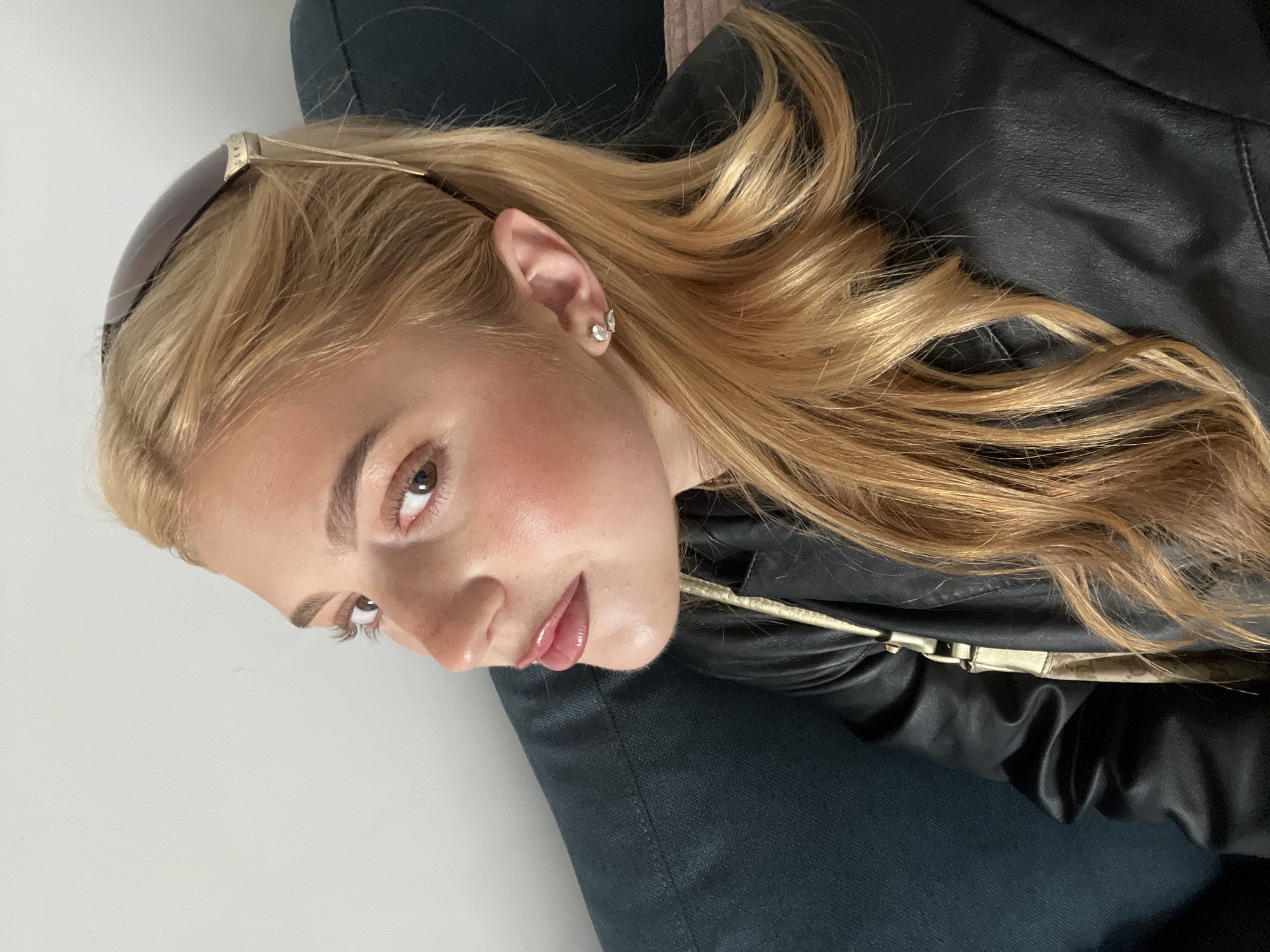 A blonde woman lies on a dark blue or black upholstered surface, possibly a sofa or chaise lounge. She wears a black leather jacket with a gold shoulder strap detail. Her hair is styled with sunglasses resting on top of her head. She has on earrings in her ear, her makeup is well-done and features rosy blush and glossy lips. The background consists of a plain white wall. The image does not contain any information that would indicate the location to be in Paris, France.
