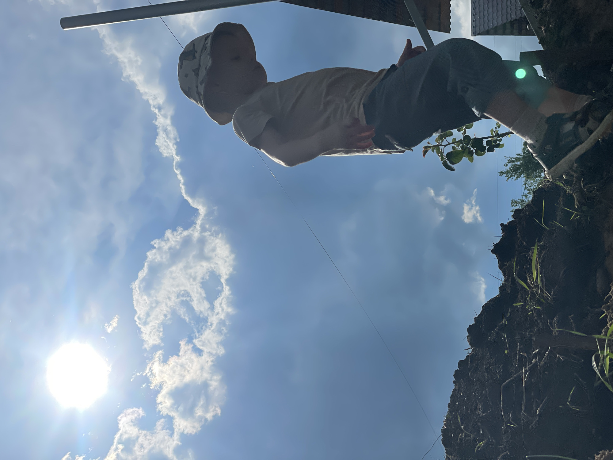 In Olkhovka, Russia, under a bright, sunlit sky with scattered white clouds, a child wearing a camouflage-patterned bucket hat and a light-colored t-shirt stands on a mound of dark soil. The child is facing downwards and to the right, with their left leg extended forward. They are wearing dark shorts and sneakers. To the right of the child, a metal pole rises vertically, and a hint of a roof structure is visible. In the foreground, the dark soil is rough and uneven, with some green grass growing through it. A thin wire stretches across the sky from left to right. The image is taken from a low angle, looking upwards.