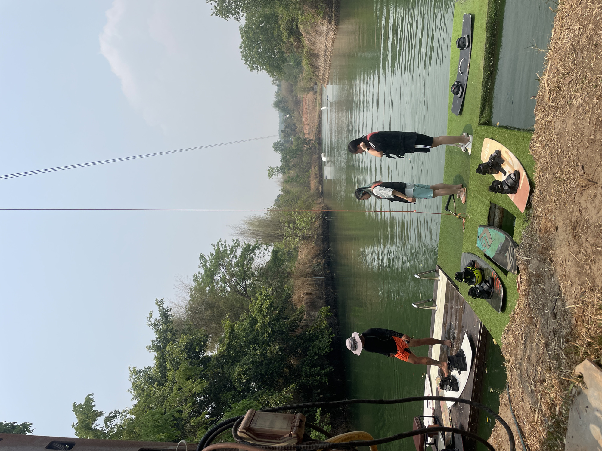 The image depicts a wakeboarding park located in Tambon Nam Phrae, Thailand. The scene shows three individuals on a dock area near a body of water, presumably a lake or pond used for wakeboarding. The individuals are wearing casual clothing and life vests, suggesting they are preparing for or have just finished wakeboarding. Wakeboards are also visible on the dock. The background consists of lush green vegetation and trees lining the banks of the water, typical of the natural environment in this region of Thailand. The sky is slightly hazy, indicating possible atmospheric conditions in the area. Overall, the scene conveys a recreational activity taking place in a natural setting in Thailand.