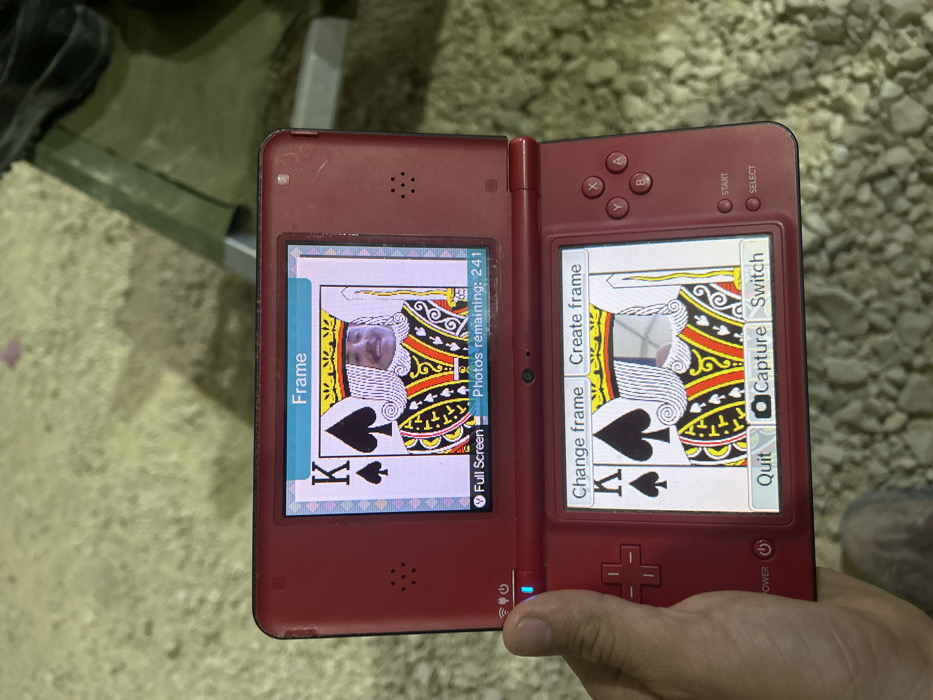 A person is holding a Nintendo DS handheld video game console in their left hand. The console's top screen displays a "Frame" of the King of Spades playing card, with a person's face superimposed onto the card's king. The bottom screen displays another "Frame" of the King of Spades playing card, but the person's face is not superimposed. The console has options for "Change frame," "Create frame," "Quit," "Capture," and "Switch." The console also has buttons for "Start" and "Select." The person is holding the console in a rough outdoor setting, with the ground beneath them being a rough, dry, pebbled surface. The location is in Tze'elim, Israel.