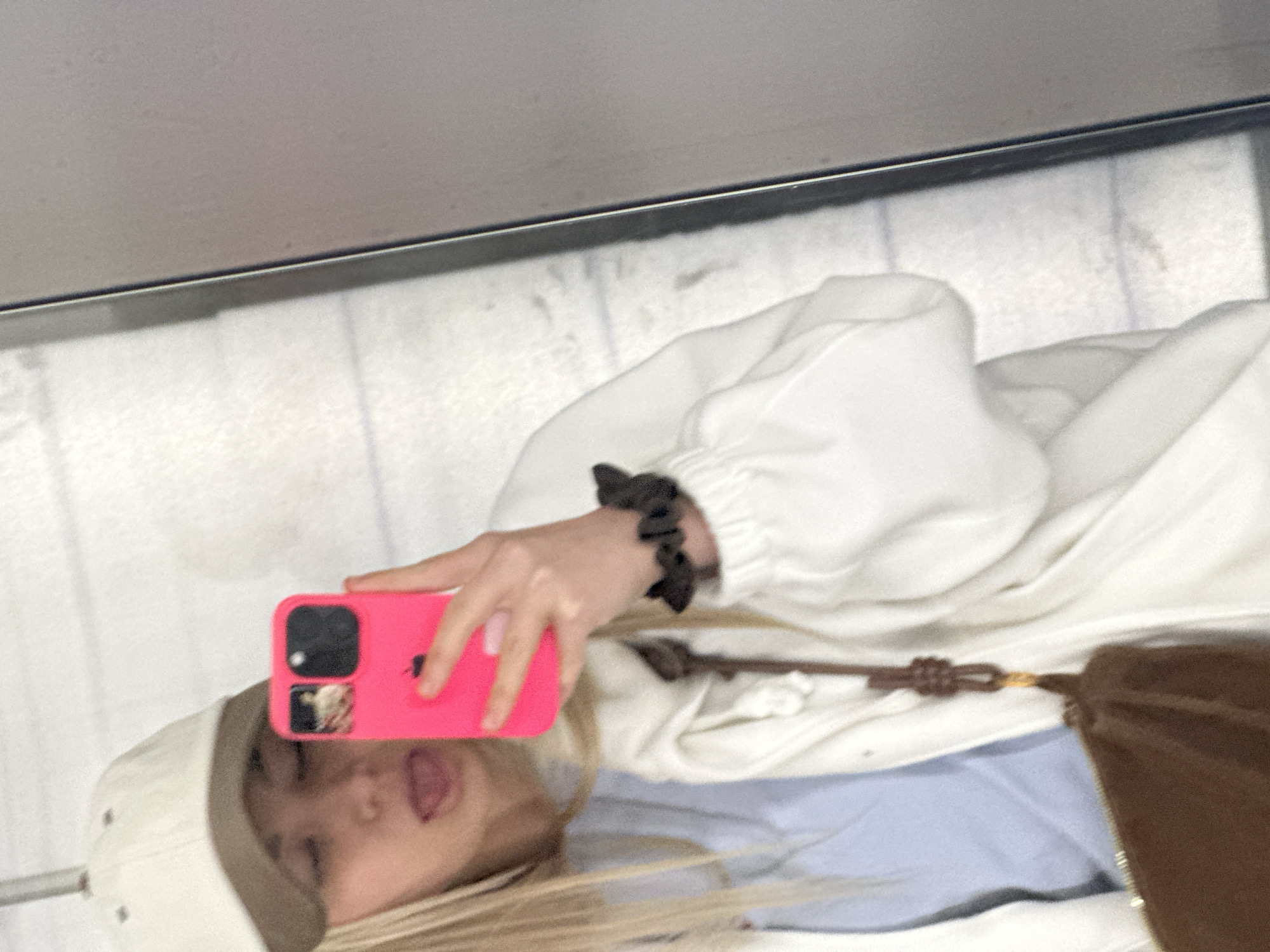 The image depicts a reflection in a glass surface, possibly a window or a glass floor. The reflection shows a person, presumably female, taking a selfie with a pink smartphone. She is wearing a white hooded sweatshirt, a light blue shirt, a beige baseball cap, and has blonde hair. Her left hand is holding the phone. A dark brown bag with a leather strap is visible near her body. The background seen through the glass appears to be a light gray or white surface, possibly tiled or concrete. The scene appears to be indoors. There is insufficient information in the image to determine the location as Budva, Montenegro.