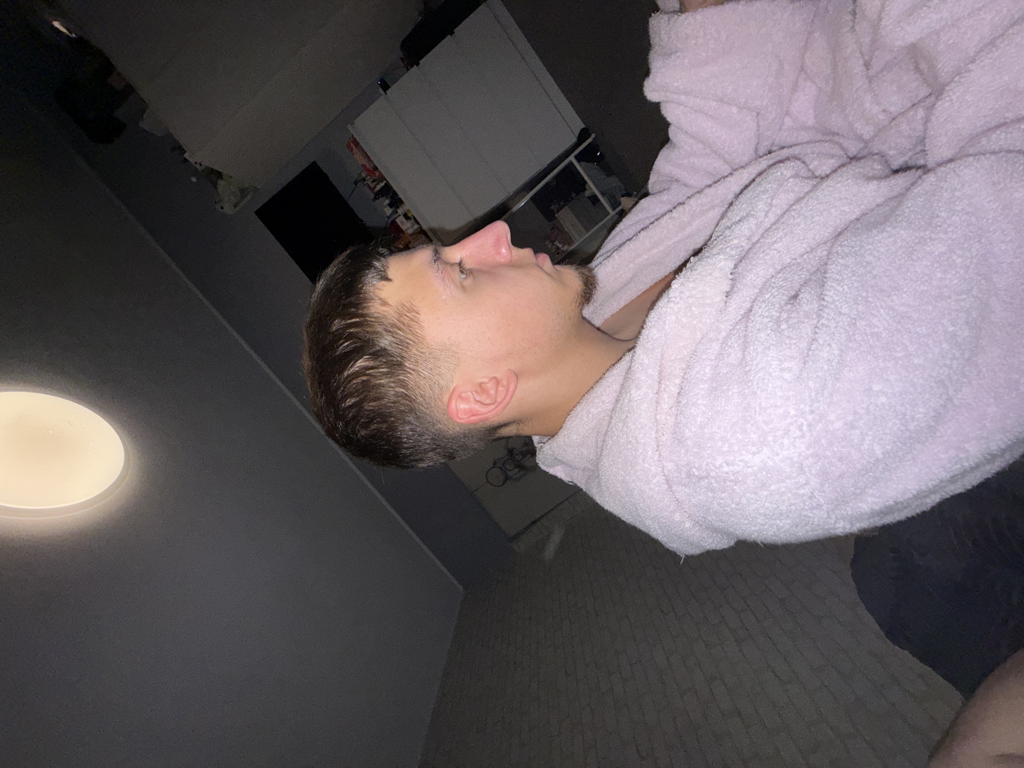 A medium, low-angle shot shows a person lying in a light pink robe indoors. The person is a young, light-skinned male with a short, dark, textured haircut, lying on his side. His head is angled to the left of the frame, with his ear visible. His face is in profile, showing a distinct nose. He has slight stubble. The robe is fluffy and covers most of his body.
The background includes a wall painted a dark shade of gray, a round ceiling light providing illumination. In the distance, there are blurry objects including a wardrobe, a shelf unit, and a television. The floor has a tile or laminate pattern. The image does not provide information to identify the location as Sofia, Bulgaria.