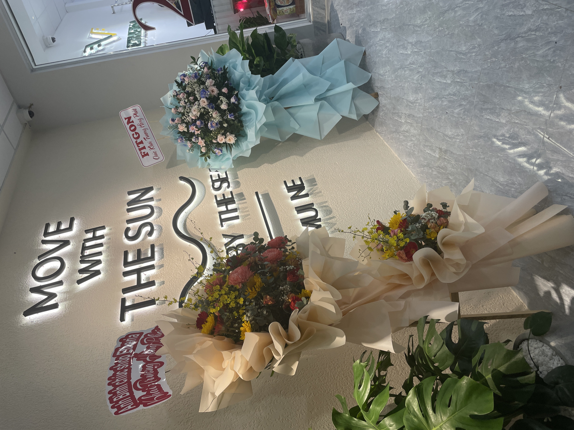 The image shows a brightly lit interior scene with a textured off-white wall as the primary background. Large, illuminated letters spelling "MOVE WITH THE SUN" are affixed to the wall. Below this, more illuminated letters spell "BY THE SEA", with a wavy line design between "SUN" and "SEA".
In the foreground, there are two large floral arrangements. The one on the left is a bouquet of pink, yellow, and red flowers, wrapped in light peach-colored paper, displayed on a wooden easel. A red sign with white lettering, reading "Anh Binh chúc shop Ebenia Khai truong hong phat", is attached to the wall to the left of this bouquet.
To the right, another floral arrangement features pink and blue flowers, wrapped in light blue paper, also displayed on a wooden easel. A red and white sign is attached to the wall next to this bouquet, reading "FITGON Chuc Khai truong hong phat". Behind this bouquet, a large potted plant with broad green leaves is visible.
The floor is tiled with a grey marbled pattern. In the background, a glass door reveals an interior with further signage, including a large red "2" and a green illuminated logo with the letter "V". Two security cameras are visible on the ceiling. The scene appears to be a store opening or celebration in Phu Thuy Ward, Vietnam.