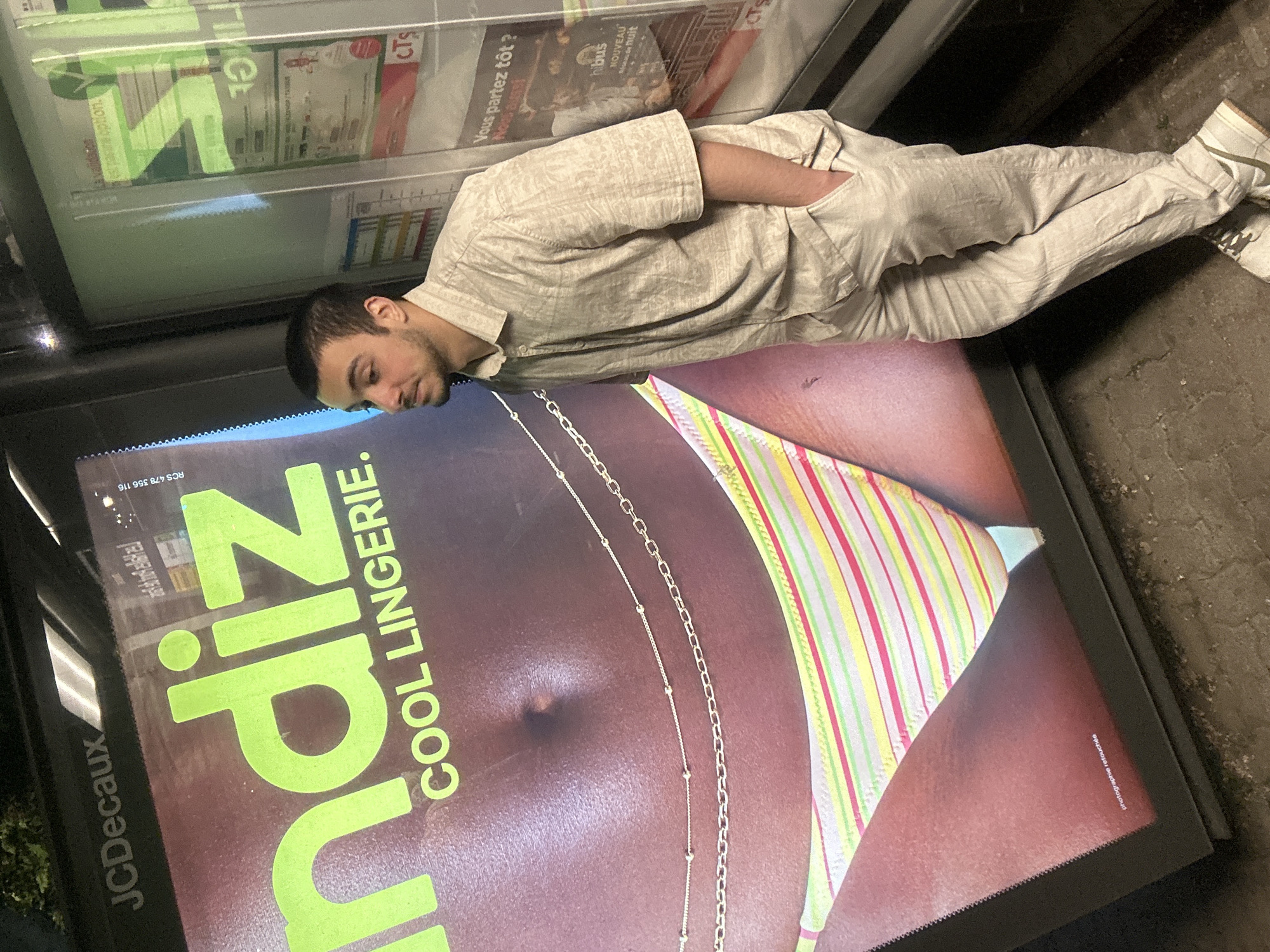 Here is a description of the image.
A man, appearing to be in his early twenties, with short dark hair and facial hair, is lying prone on top of an advertisement for a lingerie brand called "Zip", specifically "COOL LINGERIE". He is wearing a tan patterned short-sleeved shirt and tan pants with white sneakers. His left hand is in his pocket. The advertisement he is resting on features a close-up image of a woman's midsection in a striped bikini bottom with a chain around her waist. The advertisement is within a JCDecaux-branded display case, possibly a bus stop. Through the glass of the display case, other advertisements are visible. The location is in the city of Strasbourg, France.