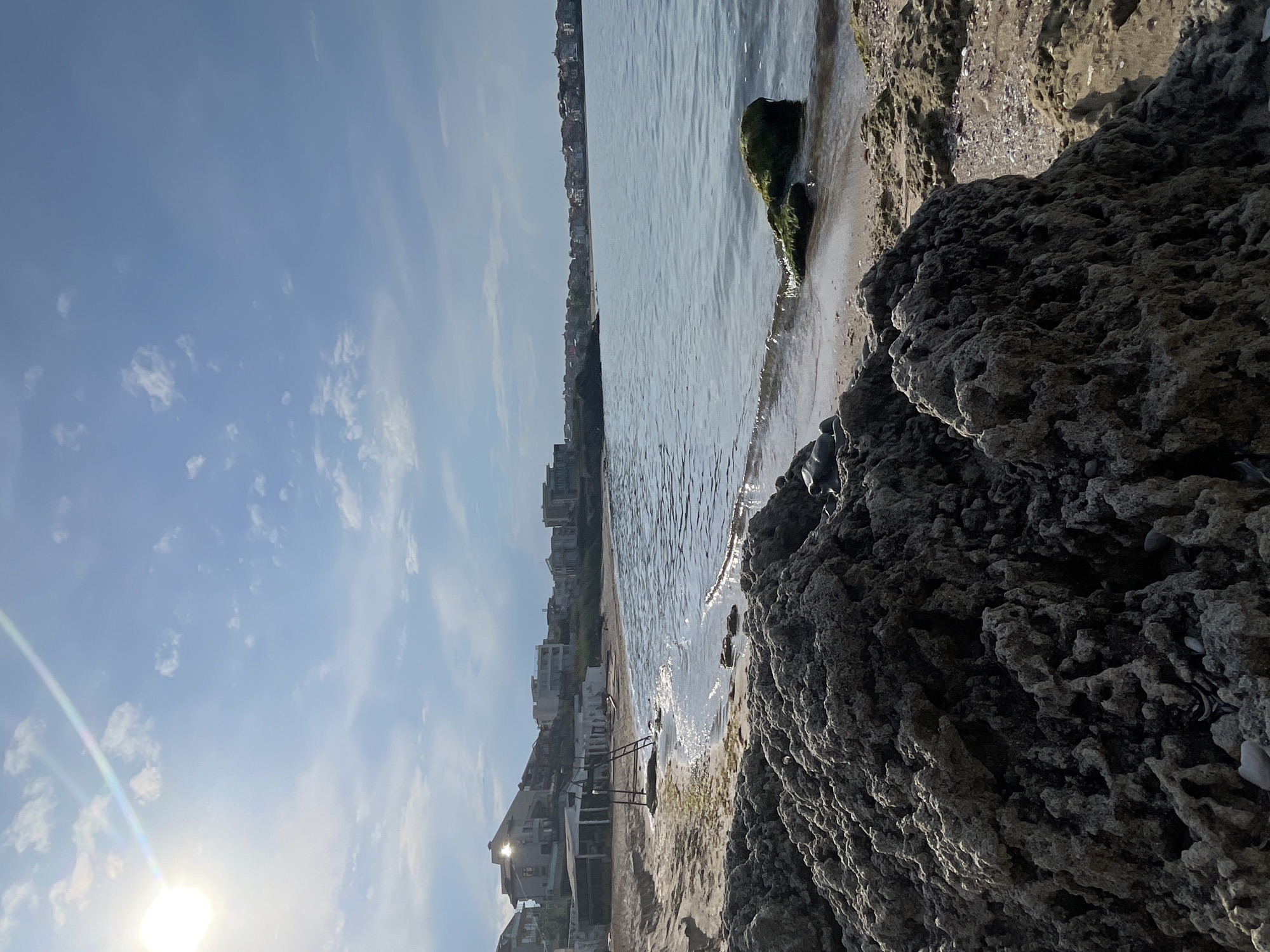 The image shows a seaside scene in Sozopol, Bulgaria. The view is from behind a rocky outcrop towards a beach. The sun is low in the sky. On the beach, there are buildings and a set of steps leading to the water. The water is calm and reflects the sunlight. The sky is blue with scattered clouds. In the distance, more buildings can be seen along the coastline. The rocky outcrop is textured and close to the camera.