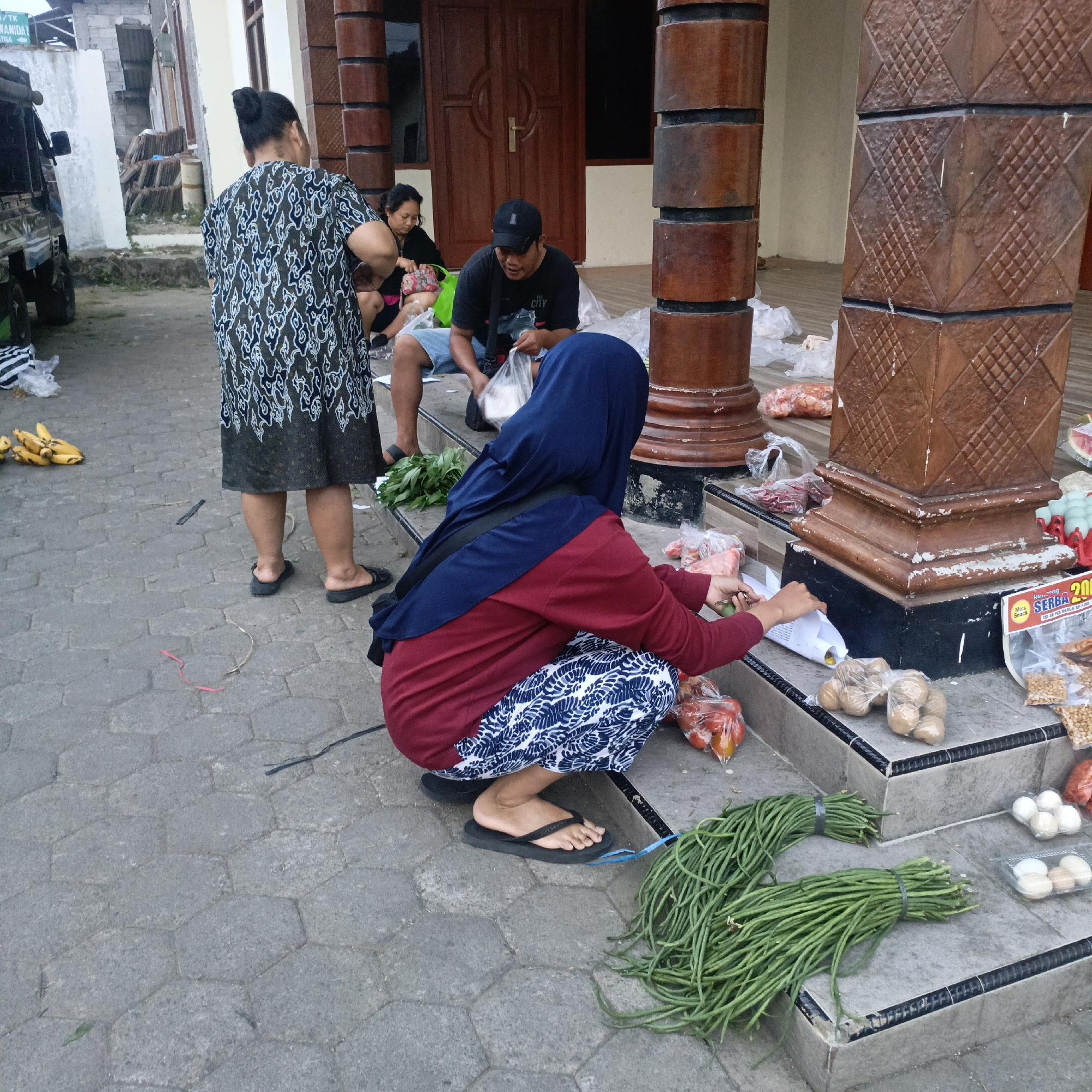 The image depicts an outdoor scene, likely a market or roadside stall, in Salatiga, Indonesia. Several individuals are present, engaged in activities related to food items.
In the foreground, a woman wearing a blue hijab and a maroon top with a patterned skirt is crouching. She is holding a piece of paper or plastic bag and appears to be interacting with food items. To her right, on a set of steps, are several bundles of long, green beans tied with rubber bands. Also on the steps are bags of potatoes and containers of eggs.
To the left of the crouching woman, a man wearing a black t-shirt and a baseball cap is sitting, holding a plastic bag and looking towards the crouching woman. Behind him, a woman in a patterned dress is standing and facing away from the camera, her back to the viewer. Further in the background, another woman is partially visible, also appearing to be engaged in some transaction or activity.
The environment is paved with hexagonal concrete blocks. In the background, there is a building with wooden doors and decorative pillars. A vehicle, possibly a truck, is parked on the left side of the image. Various plastic bags, some containing food items, are scattered around the scene, suggesting a marketplace setting. Bananas are visible on the ground to the left. A sign with red lettering, partially obscured, is also present on the steps. The overall lighting suggests it is daytime.