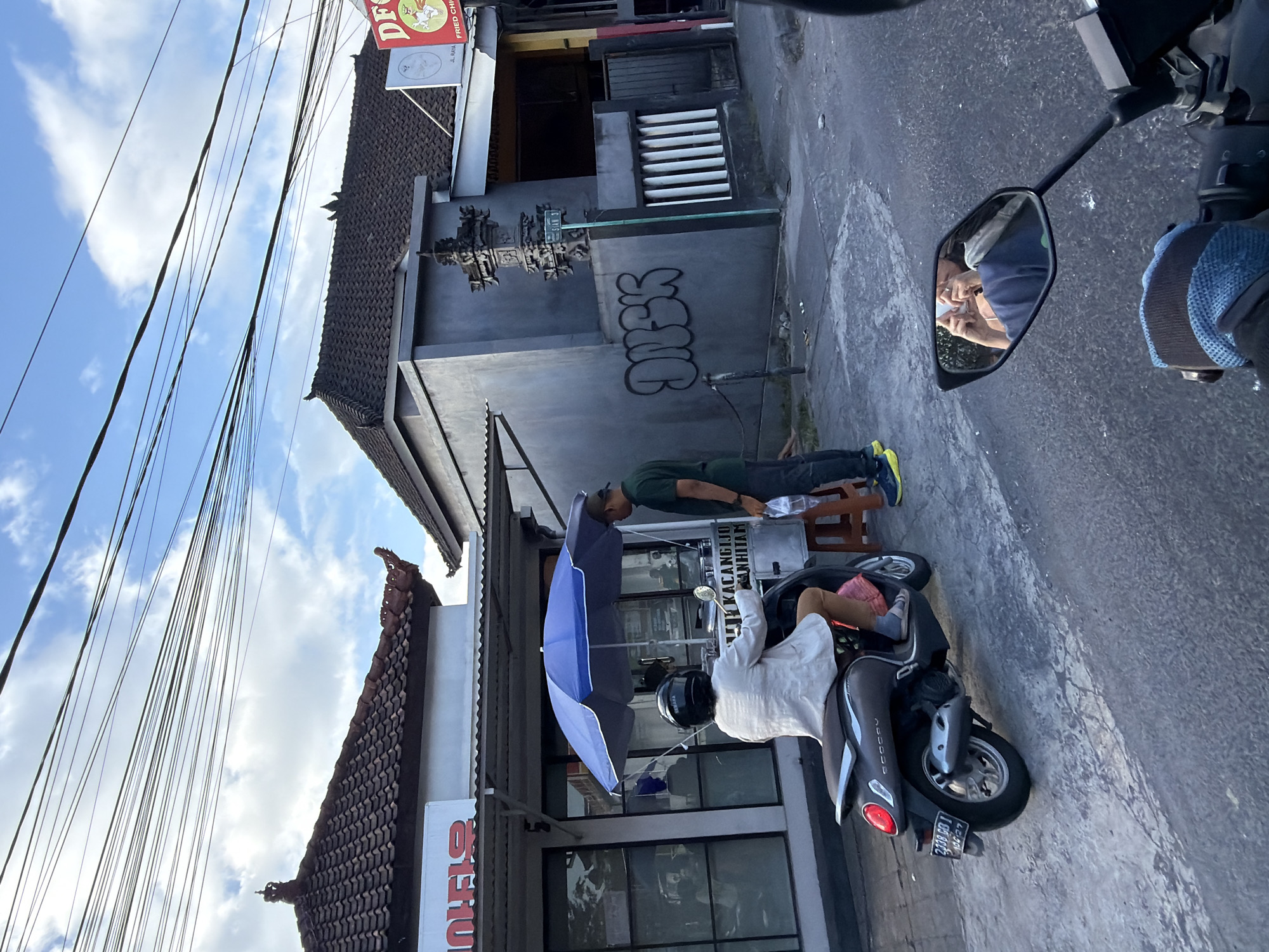 The image shows a street scene in North Kuta, Indonesia. A woman wearing a white jacket and pink pants is sitting on a dark gray scooter. A blue umbrella is open over a food cart next to the scooter. A man wearing a green shirt and dark pants is standing next to the food cart, interacting with the woman. The food cart has text on it that reads "RIJUS KACANGIJO KACANGHITAM". In the background, there are buildings with tiled roofs and a gray wall with graffiti that reads "DNSK". A sign on a pole reads "STAN S". The sky is blue with white clouds and numerous overhead wires are visible. A motorcycle mirror in the foreground reflects a person holding a phone.