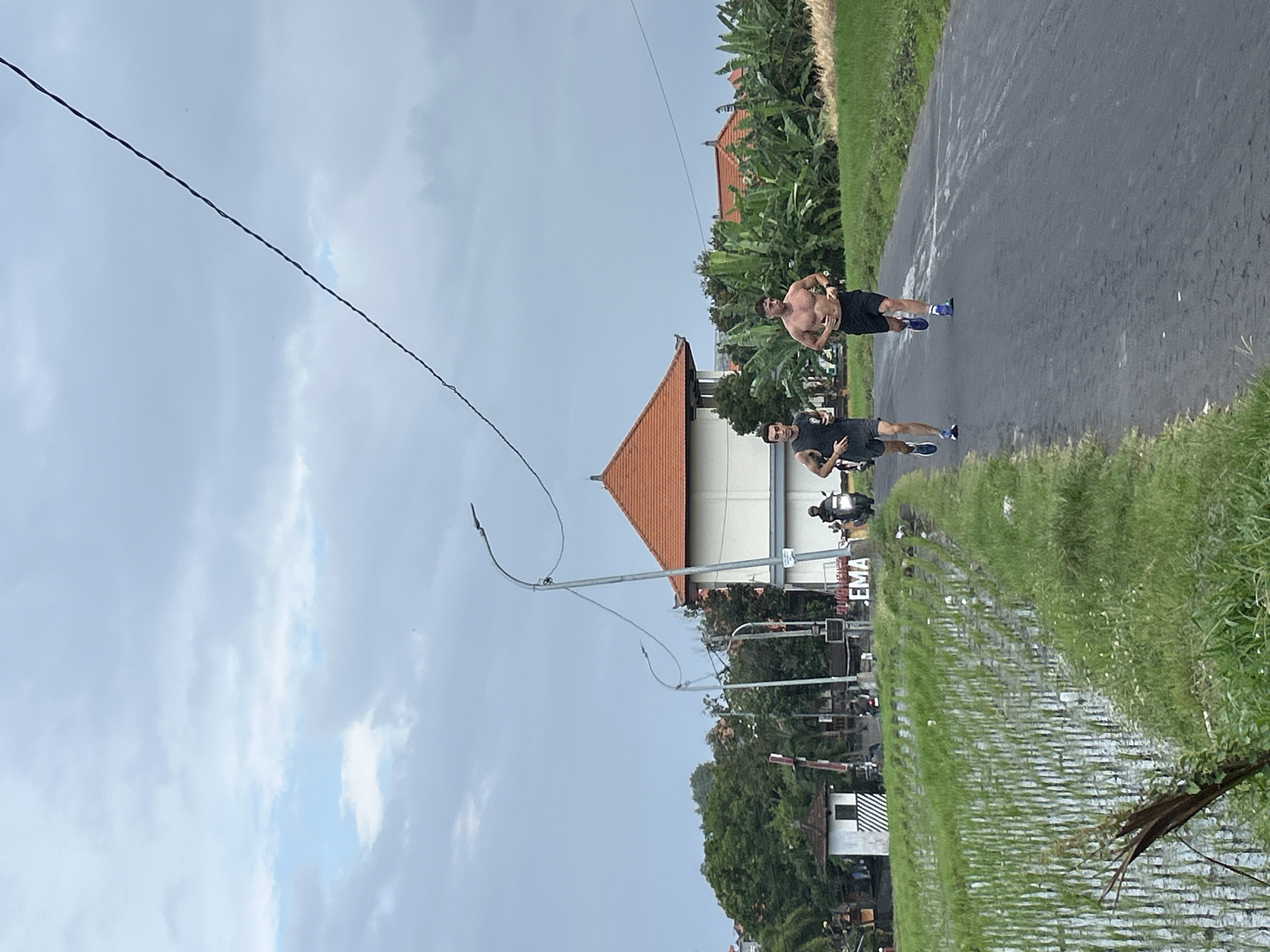 Two men are running on a paved road. The man on the left is wearing a dark t-shirt and dark shorts, while the man on the right is shirtless and wearing dark shorts. They are running on a road with a grassy shoulder and a rice paddy to the left. In the background, there is a white building with an orange roof, some palm trees, and other vegetation. There is also a utility pole with wires running across the sky. A motorcycle is visible in the distance. The sky is overcast. The image appears to have been taken in the city of Mengwi, Indonesia, as indicated by the sign in the background that reads "EMA".
