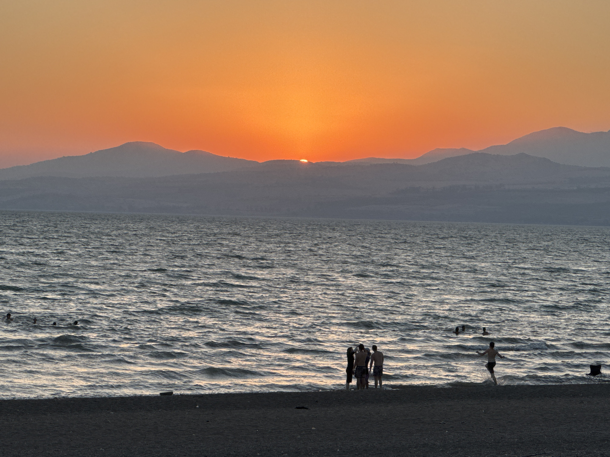 A group of people are on a beach in the Golan Regional Council, Israel, during sunset. The sun is low on the horizon, casting an orange glow across the sky and reflecting on the water. Mountains are visible in the distance, silhouetted against the vibrant sky. Some people are in the water, swimming and playing, while others are gathered on the beach. The waves are gently lapping at the shore, and the sand is dark and wet. The scene conveys a sense of relaxation and enjoyment of the natural beauty of the location.