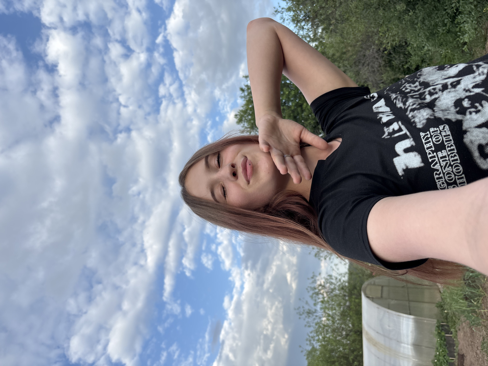 A person is looking at the camera, with their hand near their neck. They are wearing a black t-shirt that has white lettering on it. The background is a blue sky with white clouds and a greenhouse. The specific location in Фоки, Russia, cannot be determined from this image.