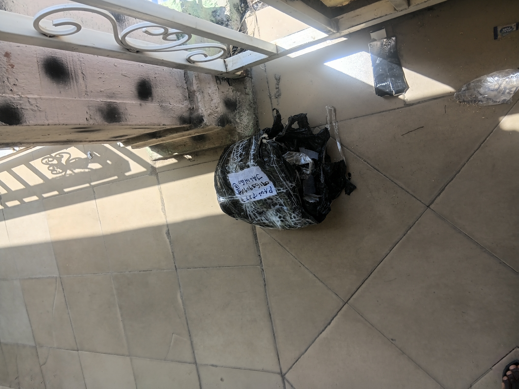 The image shows a partially wrapped package on a tiled floor, likely outside. The package is black and has a white label with writing on it. Sunlight casts shadows on the floor, indicating it is daytime. In the background, there is a white railing and a pillar with black spray paint marks. The environment appears to be an outdoor walkway or patio. The city of Jalingo, Nigeria, is mentioned, suggesting this is the location.
