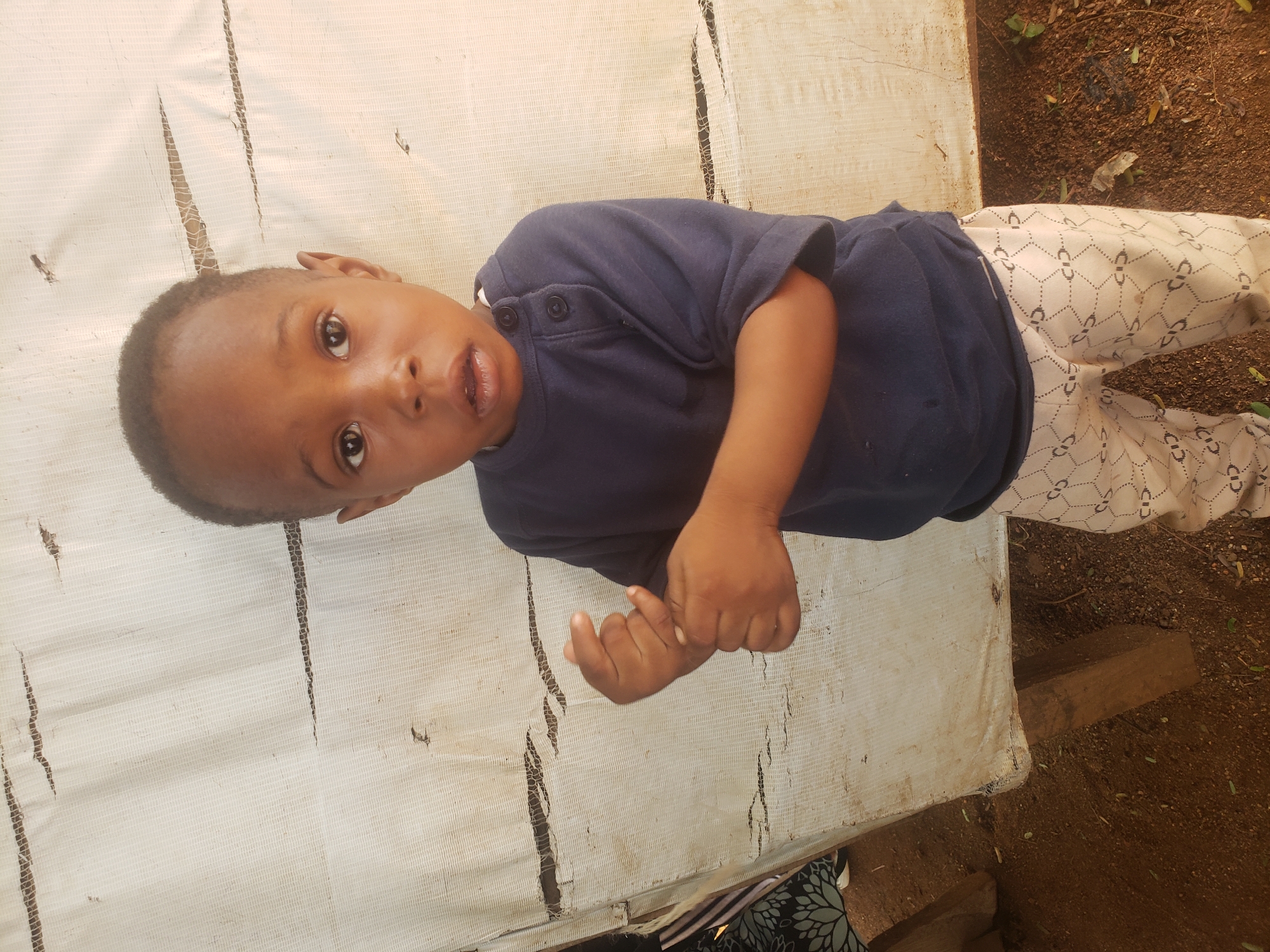 A young, dark-skinned child wearing a navy blue, short-sleeved, button-up shirt and patterned, light-colored pants lies on a tattered, white surface, possibly a sheet or tarp. The surface has visible tears and is stained. The child is looking at the camera with their mouth slightly open. The scene appears to be outdoors, with reddish-brown soil and some vegetation visible in the background. The location is specified as Adankolo, Nigeria.