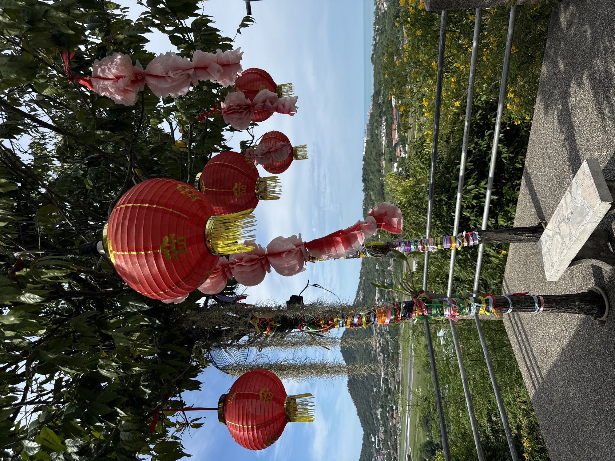 A bright, overcast day with scattered clouds highlights an outdoor viewing area with a landscape in the background. In the foreground, a stone bench sits on a textured concrete surface, framed by a metal railing. Two tree trunks, adorned with colorful ribbons and what appear to be dangling decorative strings, stand near the bench. Red lanterns with gold tassels hang from the branches of a leafy tree above. Beyond the railing, a dense green hillside slopes down towards a town, with buildings and what looks like an airport runway visible in the distance. The ocean can be seen further out, meeting the horizon. This scene is located in Ban Bang Rak, Thailand.