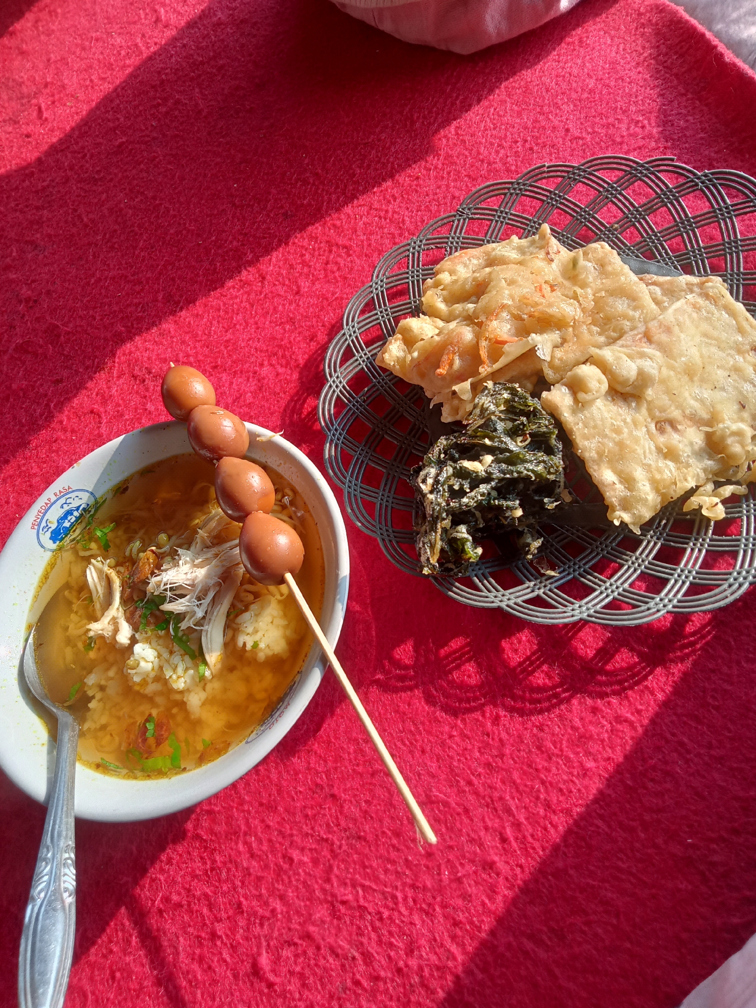 A close-up, overhead shot captures a vibrant red tablecloth as the backdrop for a traditional Indonesian meal. In the foreground, a white bowl filled with rice and shredded chicken in a savory broth, garnished with green onions, is positioned to the left. A silver spoon rests on the left side of the bowl. A wooden skewer of four small, brown, boiled eggs pierces the contents of the bowl, extending diagonally from the top of the bowl towards the right. To the right of the bowl, a woven metal dish holds two pieces of crispy fried food, likely tempeh or crackers, and a small mound of dark green, leafy vegetables. The sunlight casts distinct shadows across the tablecloth, creating a sense of depth and highlighting the textures of the food and fabric. The image appears to be taken outdoors, possibly in the city of Sruwen, Indonesia, given the presentation of the food.