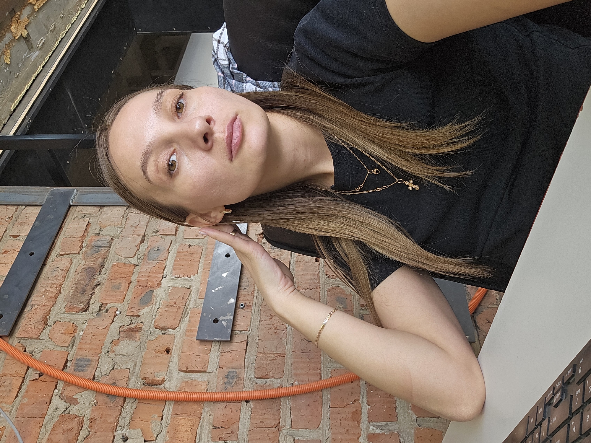 A female individual is depicted in an indoor setting, seated at a light-colored desk in the city of Chaikovsky, Russia. She has long, straight, light brown hair and a neutral facial expression, looking directly at the camera. She wears a black short-sleeved t-shirt, small gold-colored hoop earrings, a thin gold-colored bracelet on her left wrist, and two gold-colored necklaces, one with a heart charm and another with a crucifix charm. Her left hand is positioned with fingers splayed near her left ear and jawline. The background consists of an exposed red brick wall, featuring an orange corrugated conduit running vertically on the left. Dark metal structural elements and a distressed ceiling surface are visible in the upper background. A black computer keyboard is partially visible on the desk in the lower left foreground. A black and white plaid fabric object is visible behind her right shoulder.