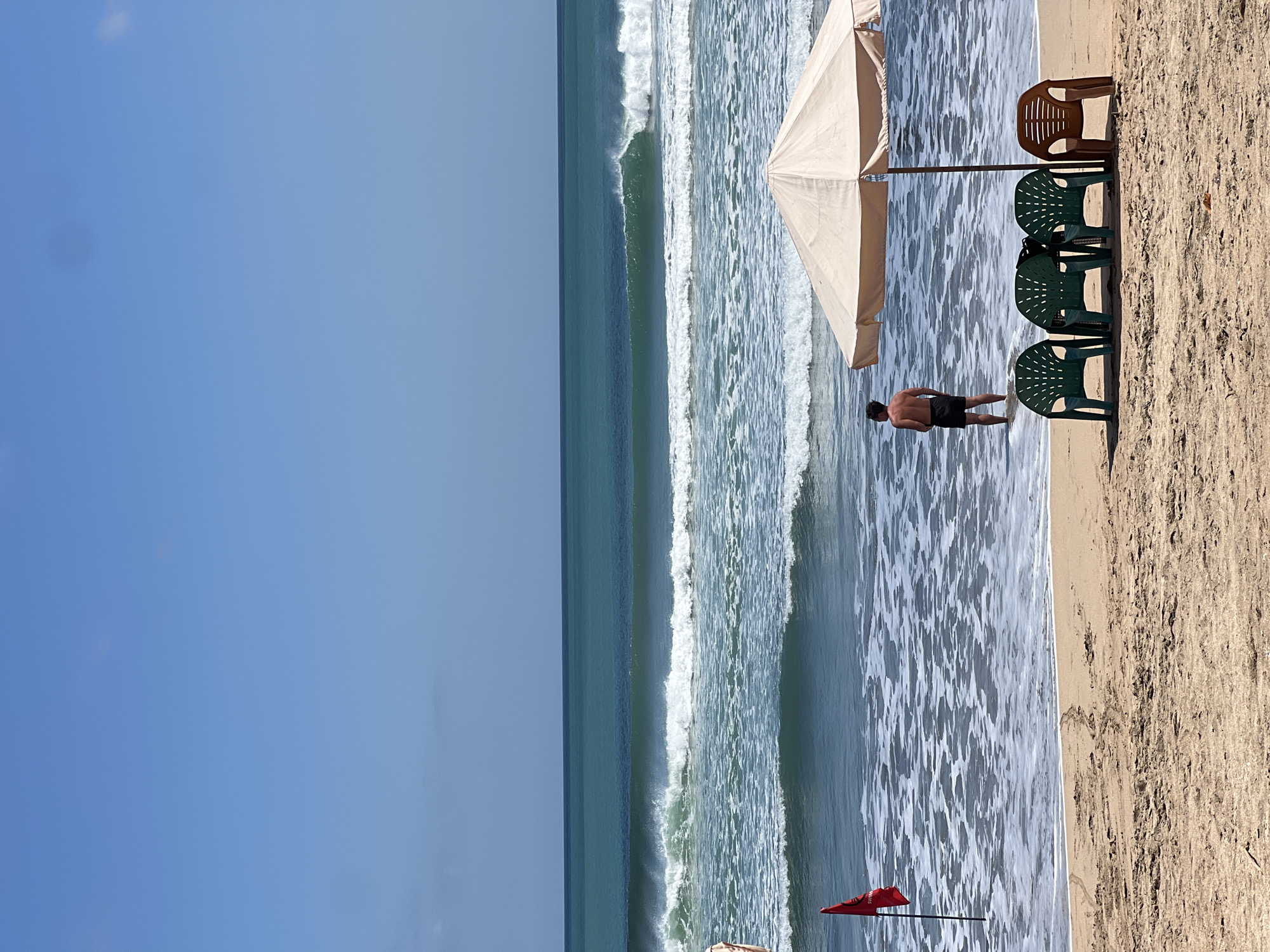 A man stands in the shallow water at Kuta Beach, Indonesia, facing the ocean. He is wearing black swim trunks. In the foreground, a beige umbrella shades three green plastic chairs and one brown plastic chair. A red flag, signaling potentially dangerous surf conditions, is planted in the sand to the left of the man. The ocean is calm with gentle waves breaking on the shore, under a clear blue sky.