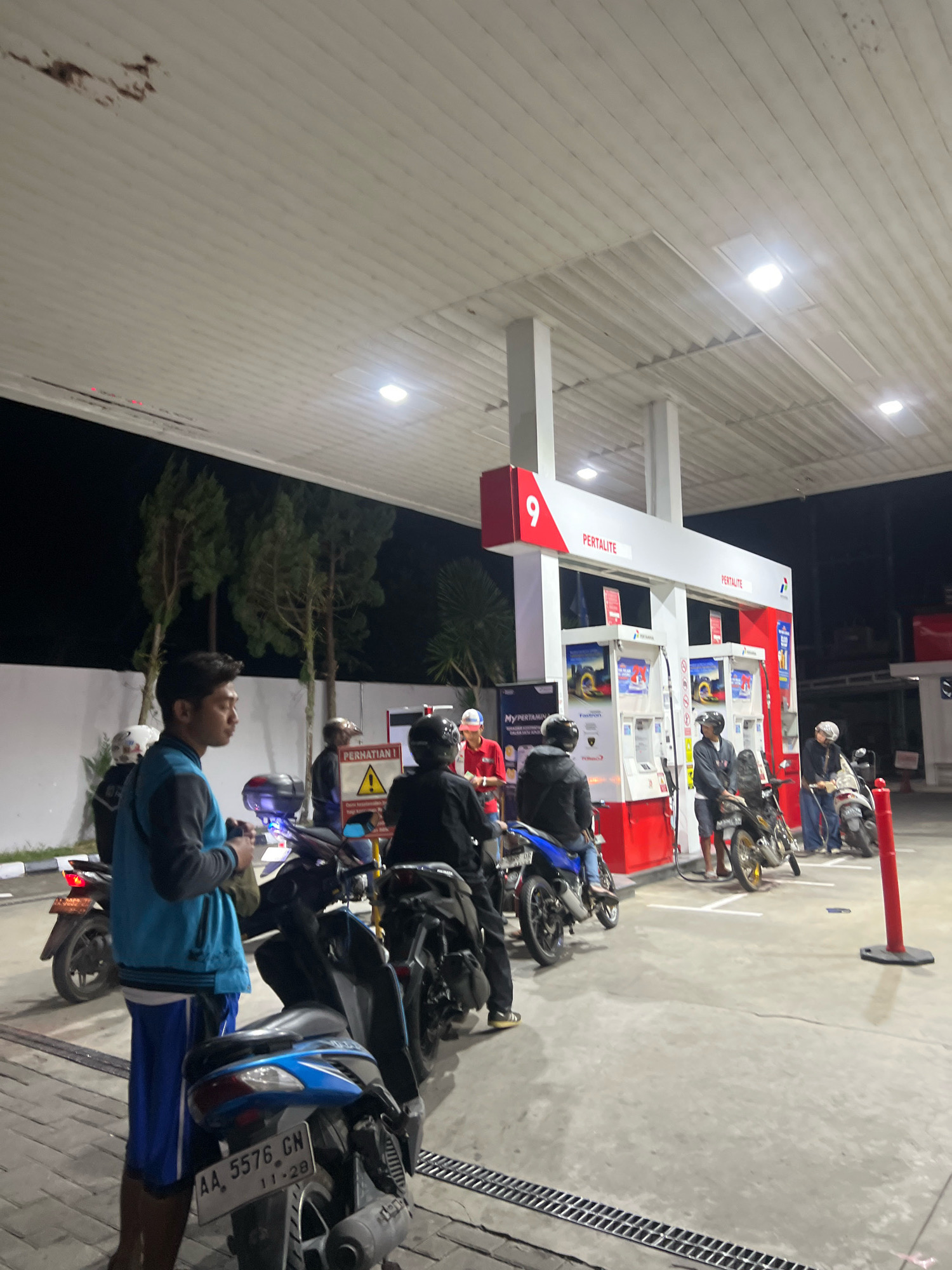 The image depicts a night scene at a Pertamina gas station in Magelang, Indonesia. Several individuals on motorcycles are waiting in line to refuel. The gas station features prominent red and white branding with the Pertalite logo visible. The lighting is provided by overhead fixtures. In the foreground, a person in a blue vest and blue shorts stands near a blue motorcycle with license plate AA 5576 GN. Other motorcycles and riders are visible further down the line. The environment includes a concrete forecourt, a white wall in the background, and trees.